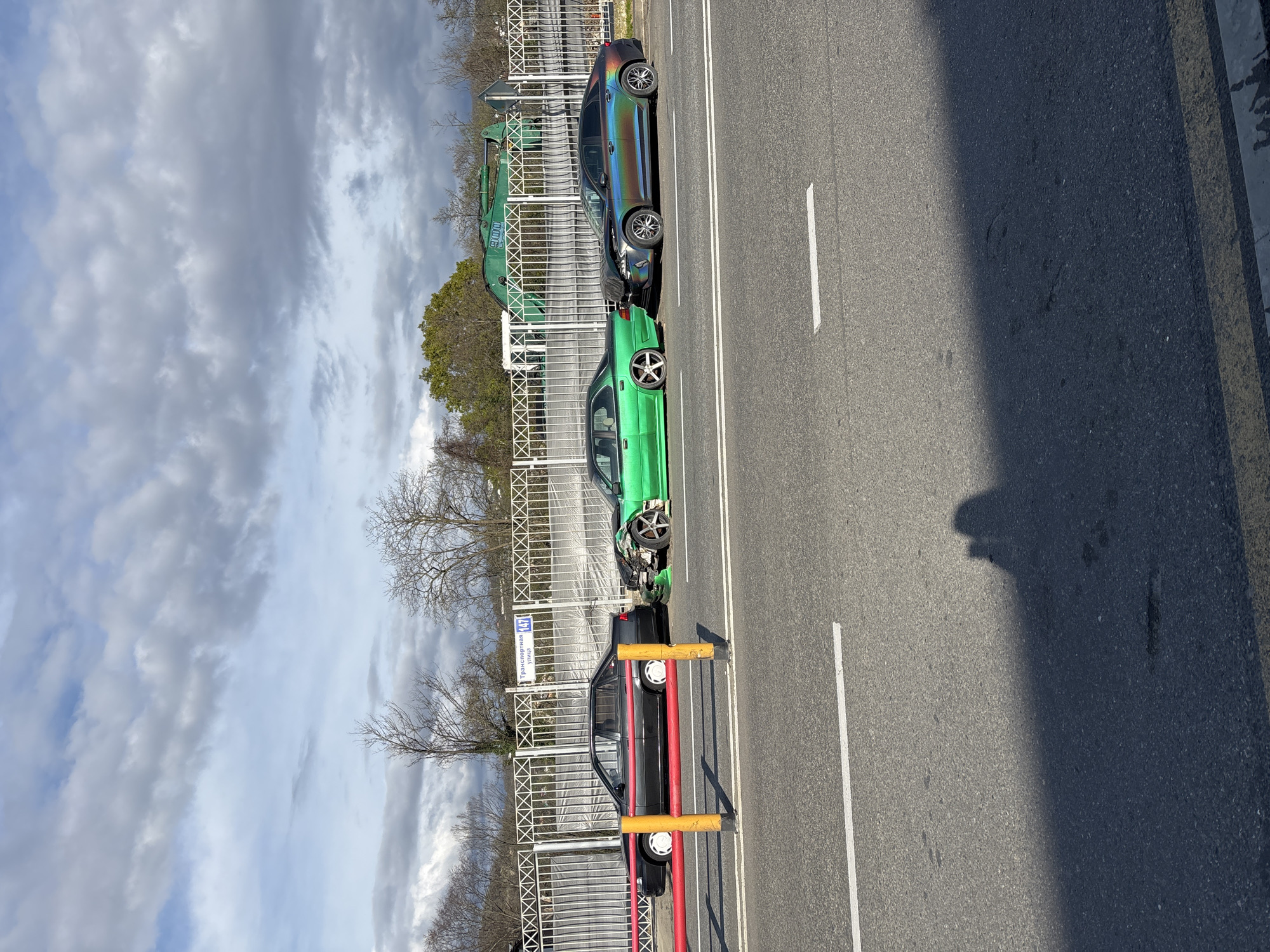 The image shows a road in what appears to be the city of Sochi, Russia. Three cars are parked along the side of the road, next to a metal fence. The closest car to the viewer is black with red accents, blocked by traffic bollards. The middle car is green and appears heavily damaged on its front driver's side. The farthest car has a rainbow iridescent paint job. A piece of construction equipment is seen behind the fence behind the rainbow car. The sky is cloudy. A road sign with Cyrillic writing on the fence near the black car reads "Транспортная улица" and "147." The road surface is asphalt, with a single white line marking the lane. The image is taken during daylight.