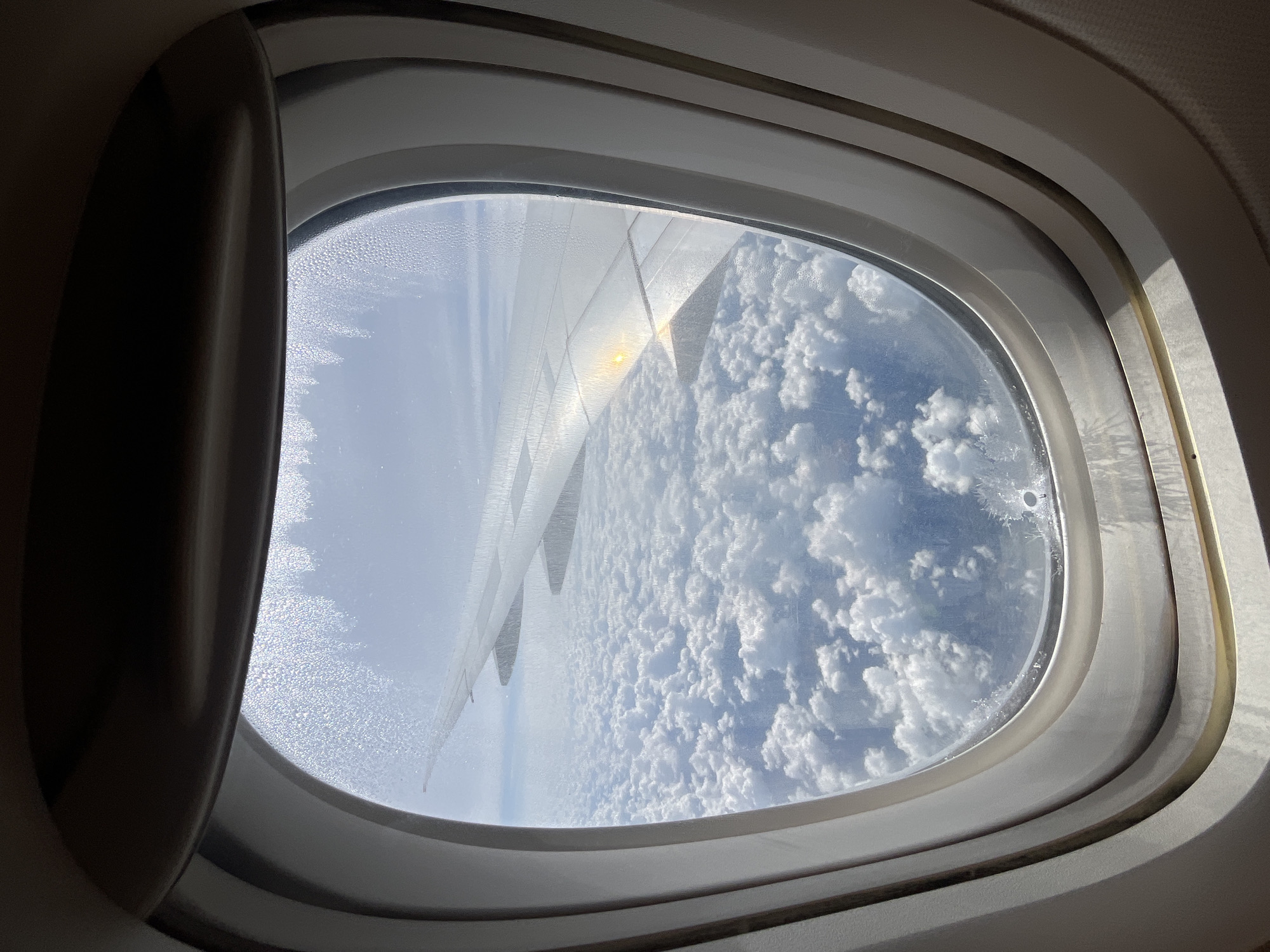 The image shows a view from an airplane window. Outside the window, a thick layer of white clouds fills the sky. An airplane wing extends into the frame from the right side, with a bright reflection from the sun visible on its surface. The window itself is framed by the interior of the airplane cabin, and condensation is visible on the glass. There are no people or identifiable structures visible in the image. The location is not discernible from the image, and therefore cannot be specified as Sukhanikha, Russia.