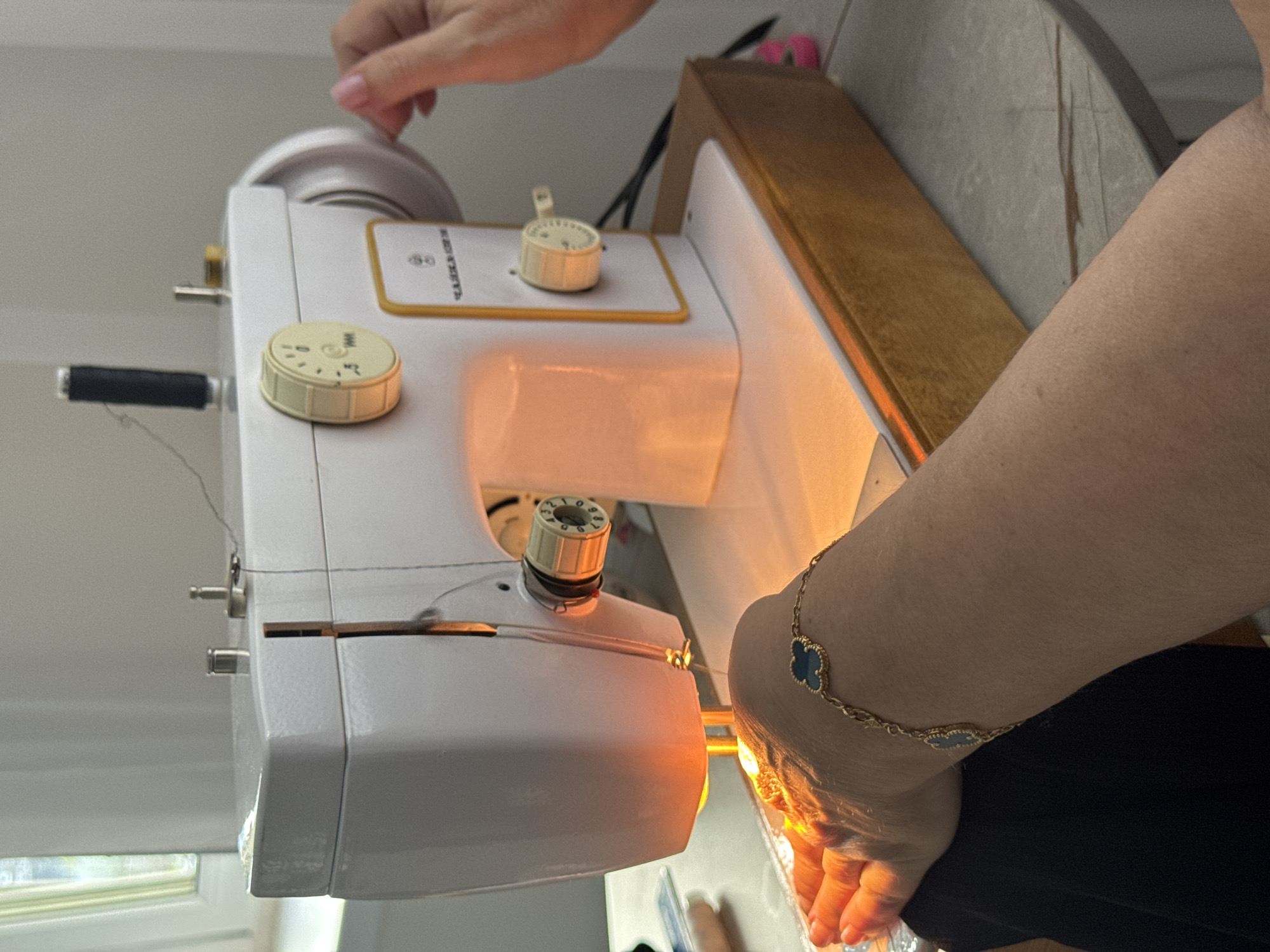 A low-angle shot captures a white sewing machine with dials and knobs, illuminated by a warm light. A hand adjusts the top wheel of the machine. Another person's arm, adorned with a gold cloverleaf bracelet, is visible near the needle area. The setting appears to be indoors, possibly in a home setting. There are no indicators of the city of Saratov, Russia.