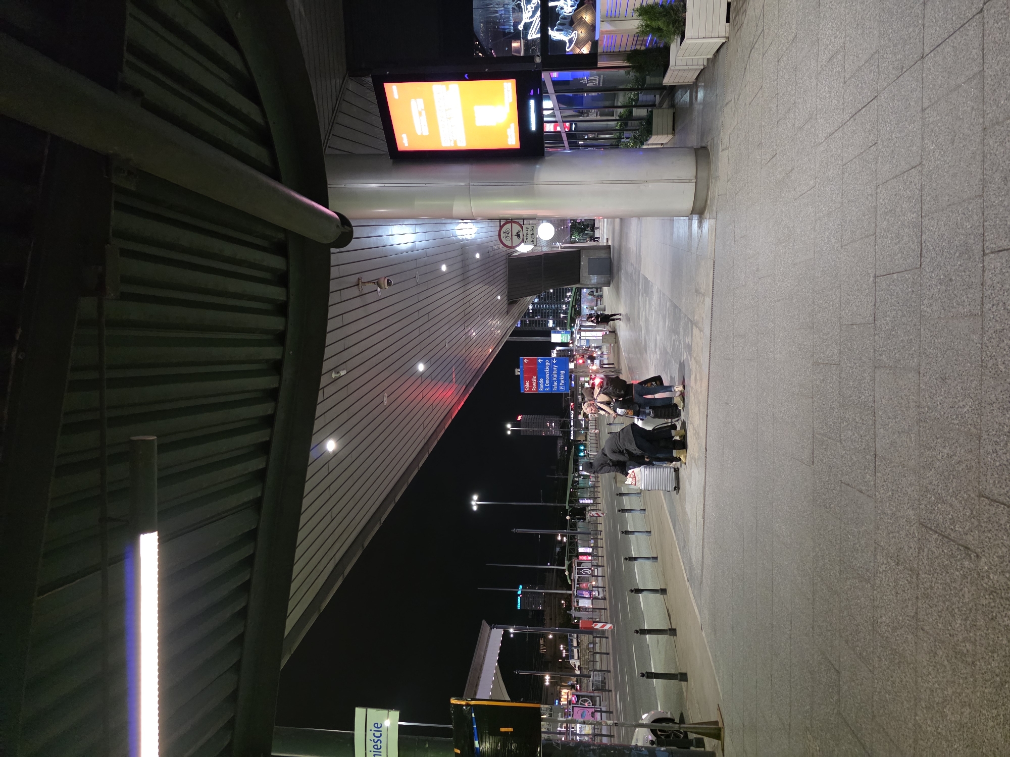 The image captures an outdoor scene at night, likely a transportation hub or a public square in Warsaw, Poland. A covered walkway with a ribbed ceiling and fluorescent lighting extends over a paved area. To the left, a green sign with white lettering reads "wnieście". Several people are gathered on the sidewalk, some with luggage, suggesting they are waiting for transportation. A silver car is parked to the far left. Across the street, there is a road with traffic lights and buildings in the background, some of which are illuminated. A large, bright orange digital display is mounted on a pillar. There are also planters with greenery visible along the side of the building. The overall atmosphere is urban and functional, characteristic of a public space in a city.