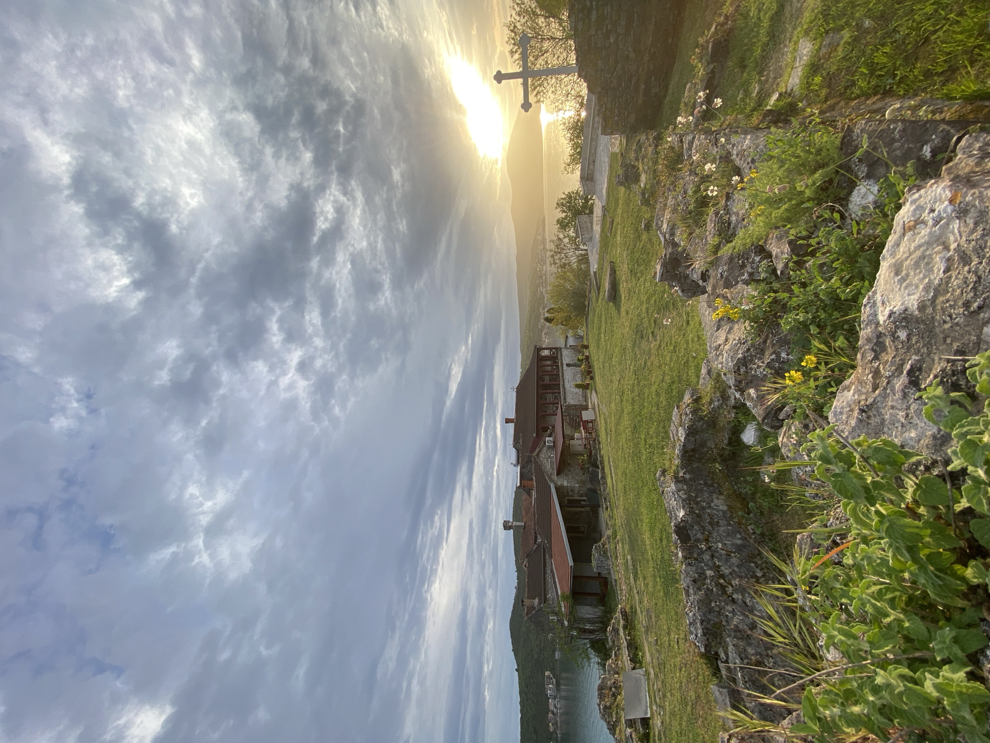The image shows a landscape in Đuraševići, Montenegro at sunset. The sky is filled with grey clouds. A cross stands on a small hill overgrown with grass and rocks. In the background are several buildings with red tile roofs and a body of water.