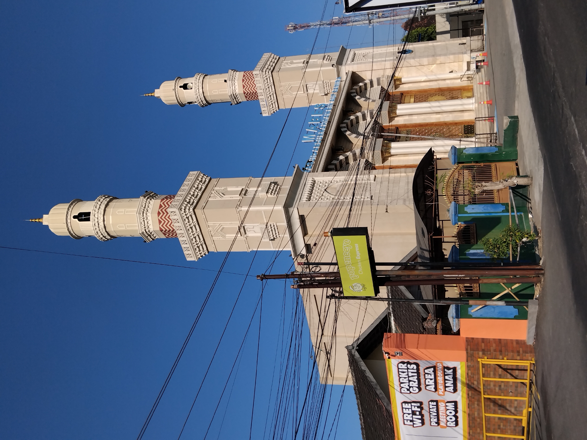 This is an exterior, eye-level, wide shot photograph of a mosque in Papahan, Indonesia, with a bright blue sky overhead. The mosque features two tall, cream-colored minarets with gold finials and decorative patterned bands in red and white. The main body of the mosque is also cream-colored with ornate detailing and large, gold-accented doors. In front of the mosque, to the left, is a billboard advertising "FREE PARKING WI-FI GRATIS PRIVATE AREA MEETING ROOM PLAYGROUND ANAK" in a colorful font on an orange and white background. A green sign for "popeneye Chicken Express" is mounted on a utility pole to the right of the billboard. The scene is a street with visible power lines crisscrossing the sky, a few orange traffic cones on the ground, and a metal fence in the foreground.