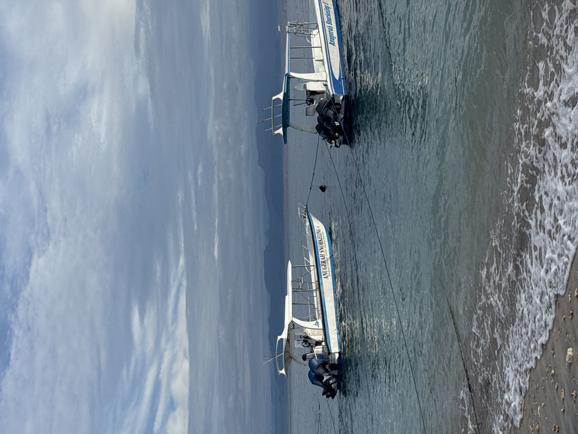 Two boats are anchored near a beach in Nusa Penida, Indonesia. The boats are white with blue trim and have outboard motors. The sky is partly cloudy. The water is clear and turquoise. The beach is sandy and rocky. The boats are labeled as "Anugerah Snorkeling."