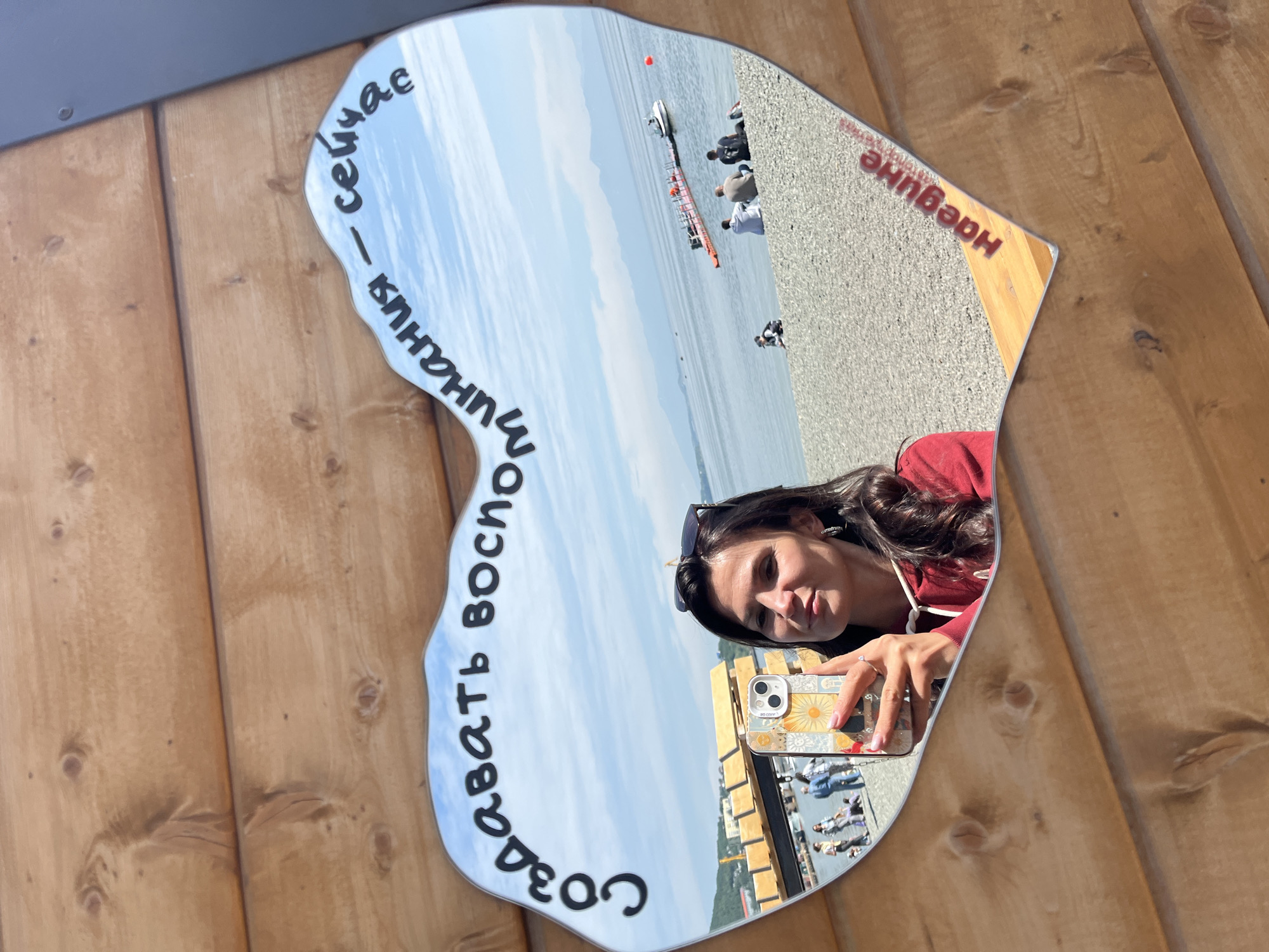 A woman takes a selfie in a heart-shaped mirror that reflects a beach scene. The mirror is mounted on a wooden wall. The reflection shows a body of water, a pebble beach with several people sitting and standing, and some boats in the water. In the foreground of the reflection, the woman is visible holding a phone. Text in Russian is visible on the mirror, with "СОЗДАВАТЬ ВОСПОМИНАНИЯ - СЕЙЧАС" ("CREATE MEMORIES - NOW") written in black along the top edge, and "НАЕДИНЕ" ("ALONE") in red on the bottom right. The location is the Avacha Bay area of Petropavlovsk-Kamchatsky, Russia.