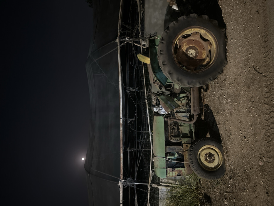 The image depicts a static scene at night in Kfar Monash, Israel, featuring a dilapidated green tractor positioned on a dirt ground surface. The tractor is severely weathered, exhibiting significant rust on its engine components, front axle, and wheel hubs, particularly on the front left wheel which is flat and appears deflated. Its body panels are faded green, and some are missing or damaged, exposing internal machinery. A yellow object, possibly part of a seat or a discarded item, is visible on the driver's side.
Behind the tractor, a large, dark mesh shade net structure, likely part of an agricultural facility or greenhouse, spans the background. This structure is supported by numerous metal poles and wires, many of which appear loose, tangled, or in disrepair. To the far left, a patch of green vegetation, possibly tall grass or weeds, grows from the ground. The sky above is dark, indicating nighttime, with a prominent, bright full moon visible in the upper-left quadrant of the frame, partially obscured by the shade net structure. No individuals are present, and no actions or interactions are depicted. The overall environment suggests a rural or agricultural setting, possibly abandoned or disused, under lunar illumination.