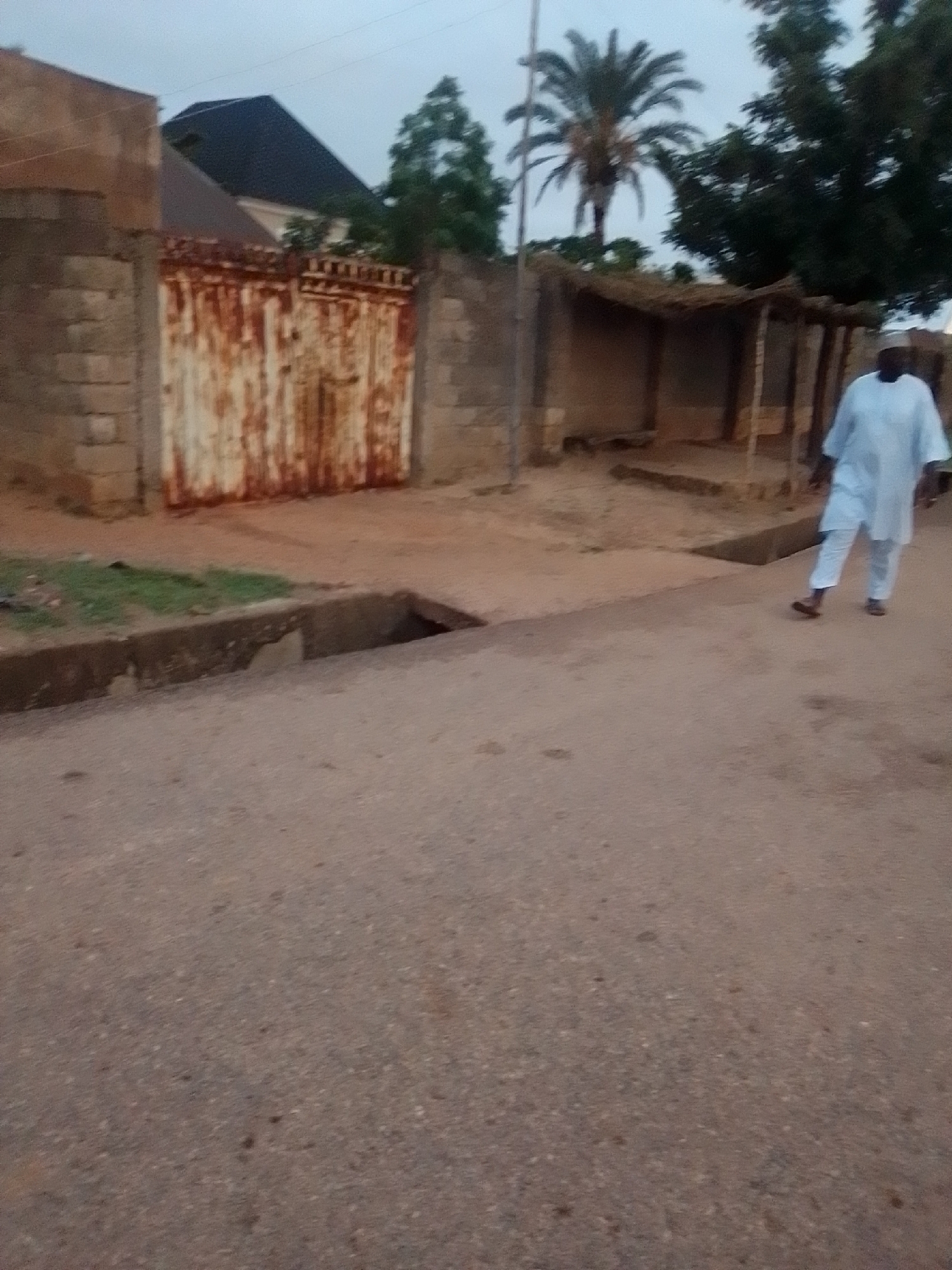 A medium shot captures a street scene in Liji Hill, Nigeria, under a muted sky. In the foreground, a paved road shows signs of wear, with a dark, open drainage ditch running along its edge. To the left, a wall made of rough-hewn blocks borders a property with a large, rusty, corrugated metal gate. Behind the wall, a dark-roofed building is visible. Further back, a cluster of thatched-roof structures stands against a backdrop of trees, including a prominent palm tree. A man, wearing a light blue tunic and trousers, walks away from the camera on the right side of the road. The environment appears to be a rural or semi-urban area, with a mixture of weathered structures and natural vegetation.