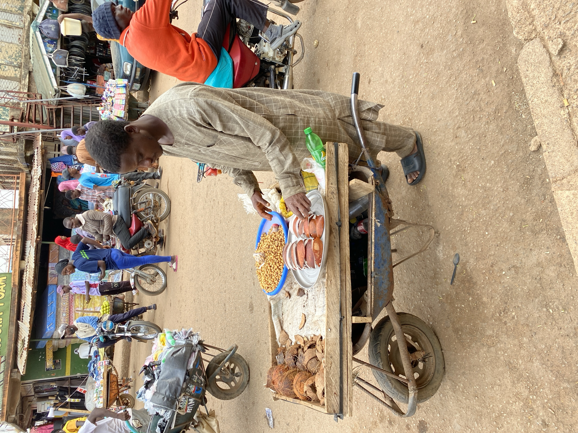 This daytime image captures a lively street scene in Bukuru, Nigeria, centered around a street vendor. The primary subject is a man dressed in a light brown, plaid-patterned traditional garment and black sandals, standing beside a rustic wooden pushcart. He appears to be attending to the items displayed on his cart.
On the cart, a blue bowl holds a generous portion of roasted chickpeas or similar legumes, alongside a white plate featuring several neatly arranged slices of what appears to be dried coconut or a related snack. Below these, the cart's lower section is filled with numerous whole and partially cracked coconuts. A green plastic bottle, possibly for a beverage, with "TEE" or "TEEL" visible on its label, also rests on the cart.
The background is bustling with activity typical of a market or busy street. Several individuals are visible, many positioned on or near motorcycles, which serve as a prominent mode of transport. One person in an orange shirt and a dark beanie is seated on a motorcycle in the upper left of the frame. Other people are standing or moving about, contributing to the vibrant atmosphere. Along the street, market stalls or small shops can be seen, with a partially visible sign that reads "FOAM." The ground is unpaved, consisting of dirt or gravel. The bright lighting indicates clear weather during the day.