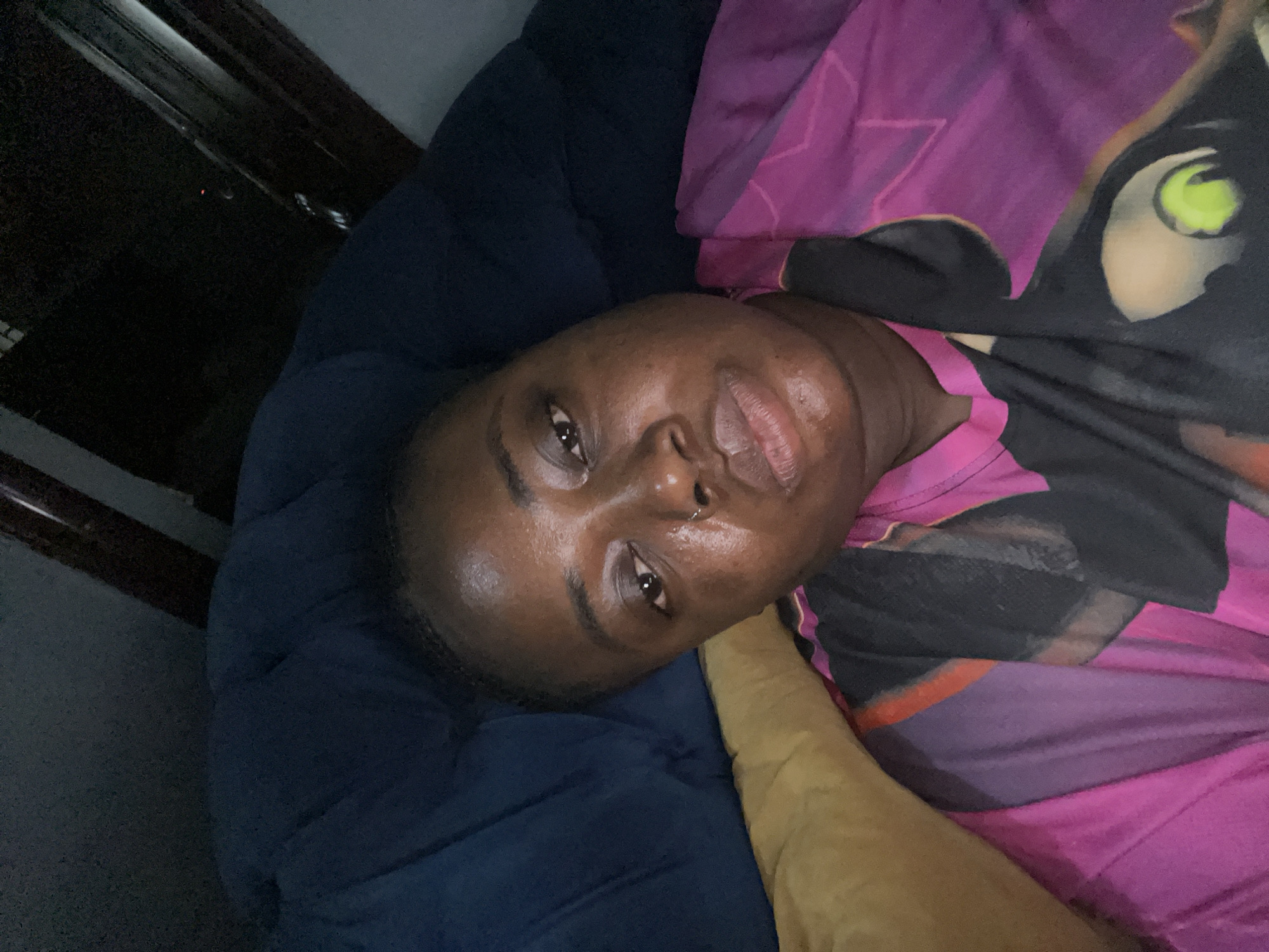 An individual lies on a blue, textured surface, possibly a sofa or bed, under pink and purple fabric with a partial cartoon-like design. The person has dark skin, close-cropped hair, a piercing in their left nostril, and is facing upward. The scene appears to be indoors. There is no information available in the image to identify the location as Kubwa, Nigeria.