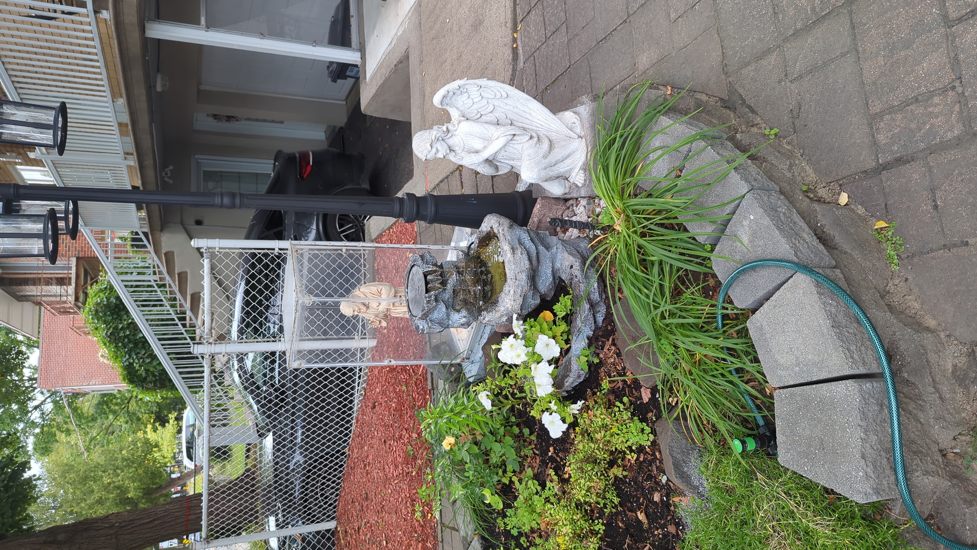 The image depicts an outdoor scene with a residential building and a garden. In the foreground, a small, tiered garden features white petunias, green foliage, and a decorative rock fountain with a statue of a monk inside a clear enclosure. To the right of the garden is a statue of a winged angel kneeling with its hands clasped. A black lamppost stands behind the garden and angel statues. A green garden hose is coiled near the edge of the garden bed, connected to a spigot. In the background, a chain-link fence encloses a driveway with dark-colored vehicles. The building behind the fence has multiple levels with balconies and an external staircase. The scene appears to be in a residential area of Montreal, Canada, possibly in the summer or fall, given the presence of green foliage and flowers.