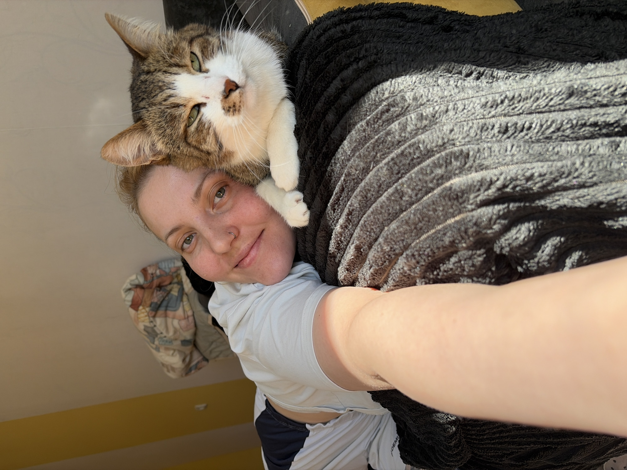 A woman takes a selfie indoors with her cat perched on her shoulder. The woman is fair-skinned with light brown hair pulled back. She is wearing a light-colored t-shirt and has a nose ring. The cat is a tabby with white markings and green eyes. They are both lying on a dark-colored textured blanket. The background shows a ceiling and part of a wall. The location is indicated to be Chaikovsky, Russia.