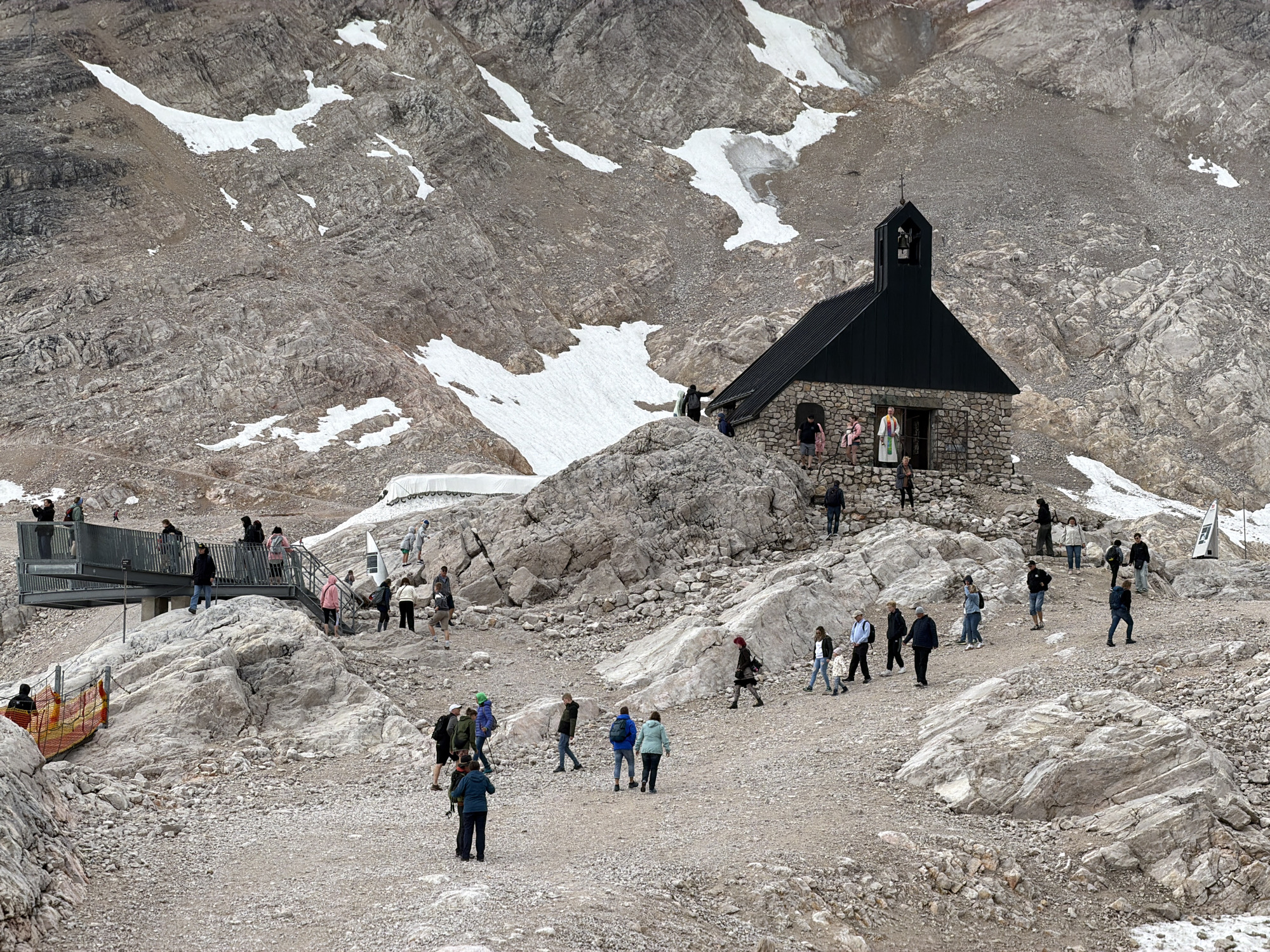 The image depicts a rocky, mountainous terrain with patches of snow, located near the summit of the Zugspitze, the highest peak in Germany, which is a popular tourist destination accessible from Garmisch-Partenkirchen. A small stone building with a dark, sloped roof and a bell tower is situated prominently in the middle ground. Several people, dressed in casual clothing, are scattered around the building and on pathways, engaging in various activities such as walking, standing, and observing the surroundings. A metal walkway or viewing platform is visible on the left side of the image, also occupied by a few individuals. The overall environment is rugged and mountainous, characteristic of high-altitude locations.