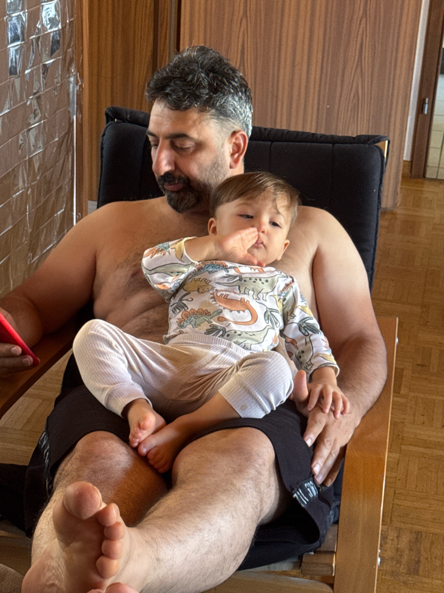 The image shows a shirtless man sitting in a chair with a baby on his lap. The man has dark hair and a beard and is holding a red object, possibly a phone, in his left hand. The baby is wearing a white long-sleeved shirt with a dinosaur print and light-colored pants. The baby's right hand is near its mouth, and its left hand is resting on the man's hand. The background appears to be a room with wooden walls and flooring. The image does not contain any specific details or landmarks that would allow for its location to be pinpointed to Überherrn, Germany.