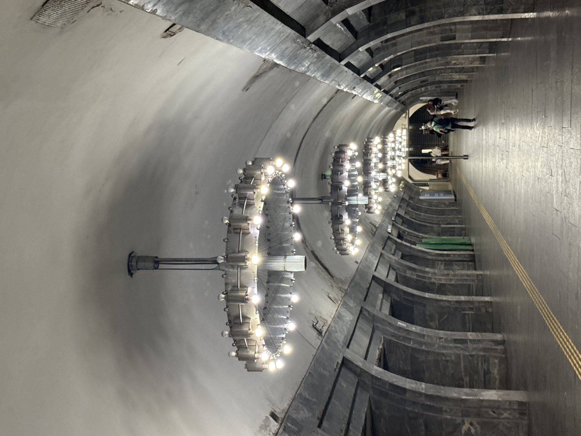 This is a photograph of the Kyiv Metro, specifically the Zoloti Vorota (Golden Gate) station. The image depicts a long, arched hallway with a high, vaulted ceiling. Several large, circular chandeliers with multiple light bulbs hang from the ceiling, illuminating the space. The walls are constructed of dark, polished stone with a repetitive, arched architectural motif. A yellow tactile paving line runs along the edge of the platform. In the distance, several people are visible walking along the platform. One person, wearing a green shirt and a backpack, is walking away from the camera towards the distant figures. The station appears to be clean and well-maintained, with a somber and grand aesthetic.
