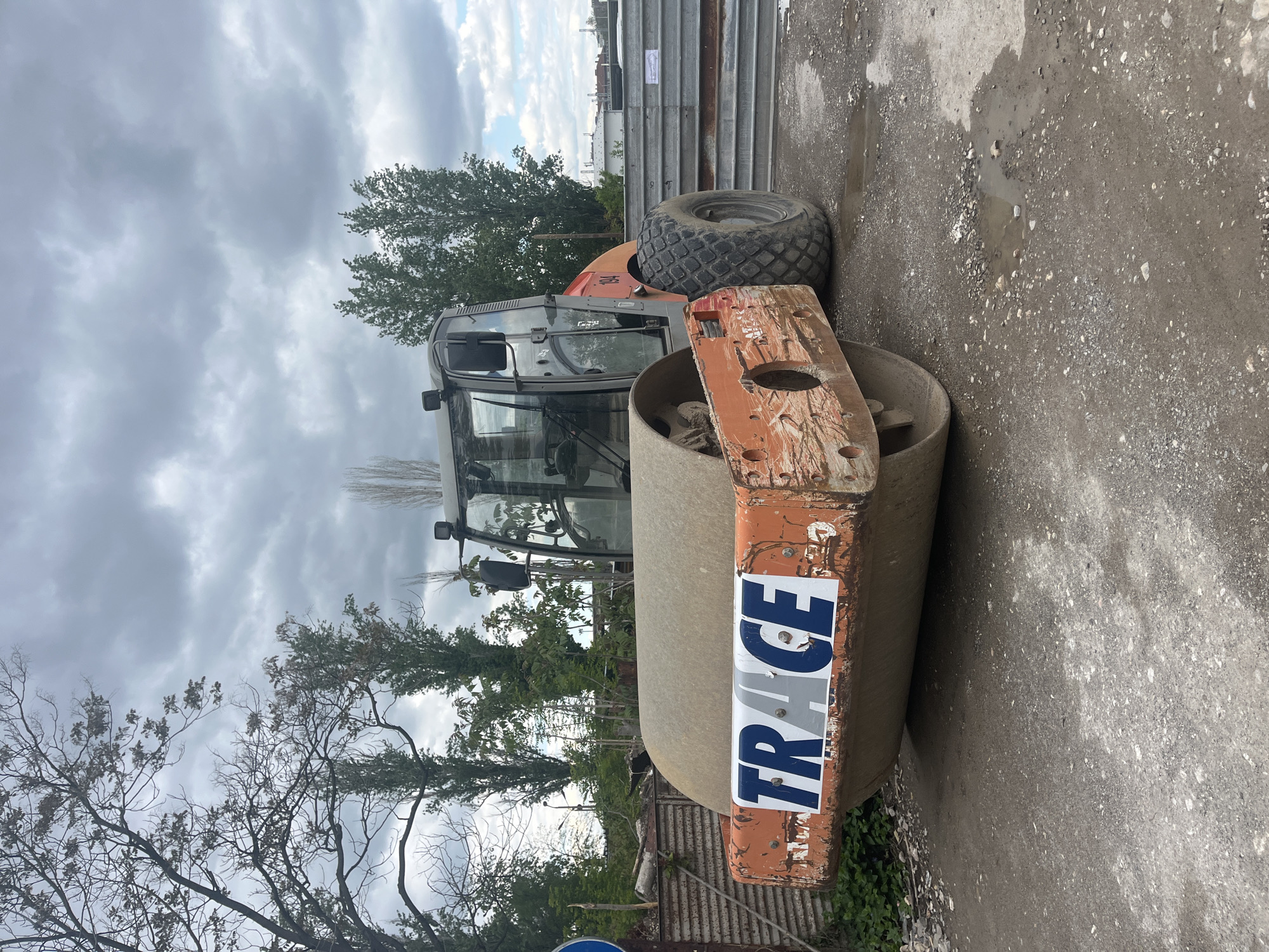 The image shows a road roller with the word "TRACE" written vertically on its side. The roller is parked on a gravelly surface that also has some wet spots. The background includes some trees and a cloudy sky. The image was taken in an urban area of Plovdiv, Bulgaria.