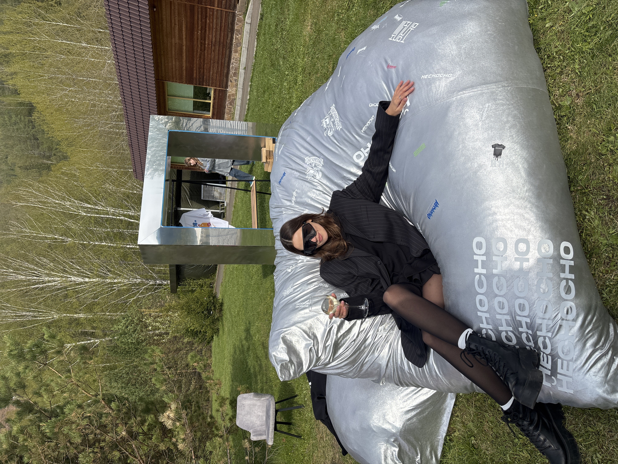 Here is a description of the image:
A young woman in a black blazer, dress, black tights, and black boots reclines on a large silver inflatable bag in a grassy outdoor setting. She is wearing sunglasses and holding a glass of what appears to be wine. The bag has the word "HECHOCHO" printed multiple times on its surface.
In the background, there's a wooden building with a brown roof and some trees, including birches. A shiny, reflective structure is visible near the woman and the building. There's also a gray chair near the bag. The setting appears to be a grassy area near a building or structure with reflective surfaces. The exact location cannot be determined as the image does not have any context clues to identify a street name or specific landmarks. There is no information provided to suggest that this is in Krasnoyarsk, Russia.