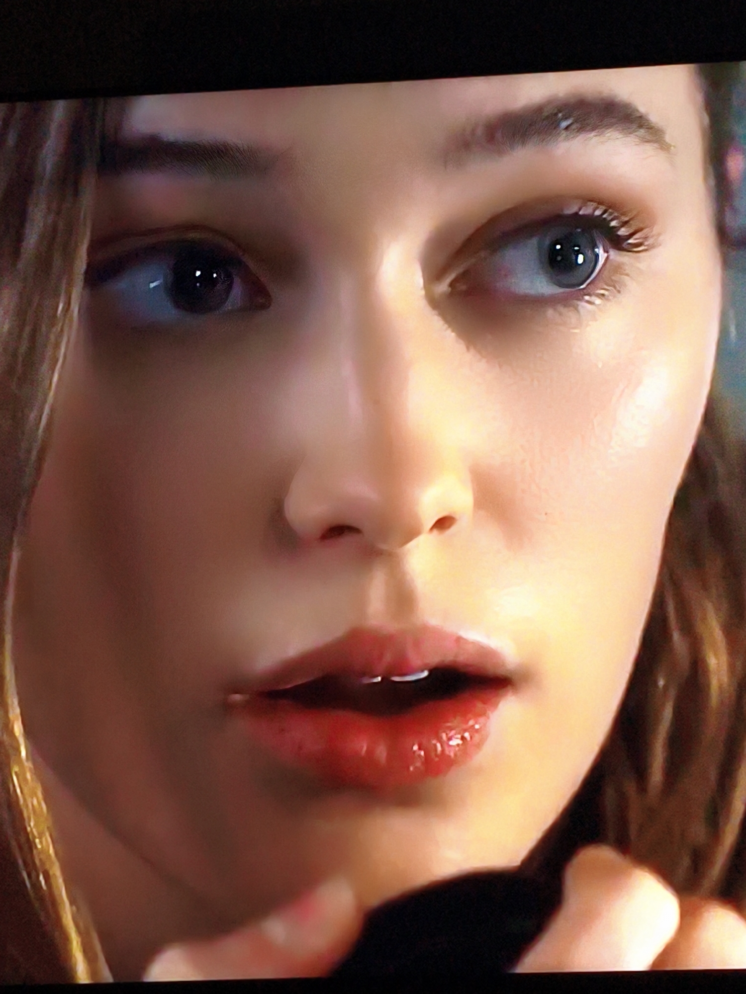 A close-up shot focuses on the face of a young woman with brown hair. Her eyes are wide, looking slightly to her right, with dark pupils and light irises. Her eyebrows are neatly shaped. Her lips are full and painted with red lipstick, slightly parted, revealing her teeth. The skin on her face appears smooth and illuminated, with some subtle highlighting on her cheekbones. The background is dark and out of focus, making the woman's face the sole subject of the image. There is no indication of the city of Vladivostok, Russia, or any specific environmental context.