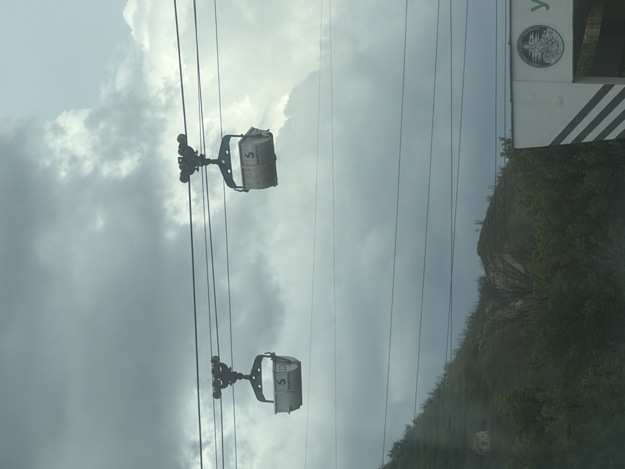 The image shows two cable cars hanging from cables against a cloudy sky. The cable cars are branded with the letter 'S' and the number '229'. In the background, there's a mountainside covered in greenery. Part of a sign or building with a circular emblem can be seen on the right. It's Kipra, Bulgaria.