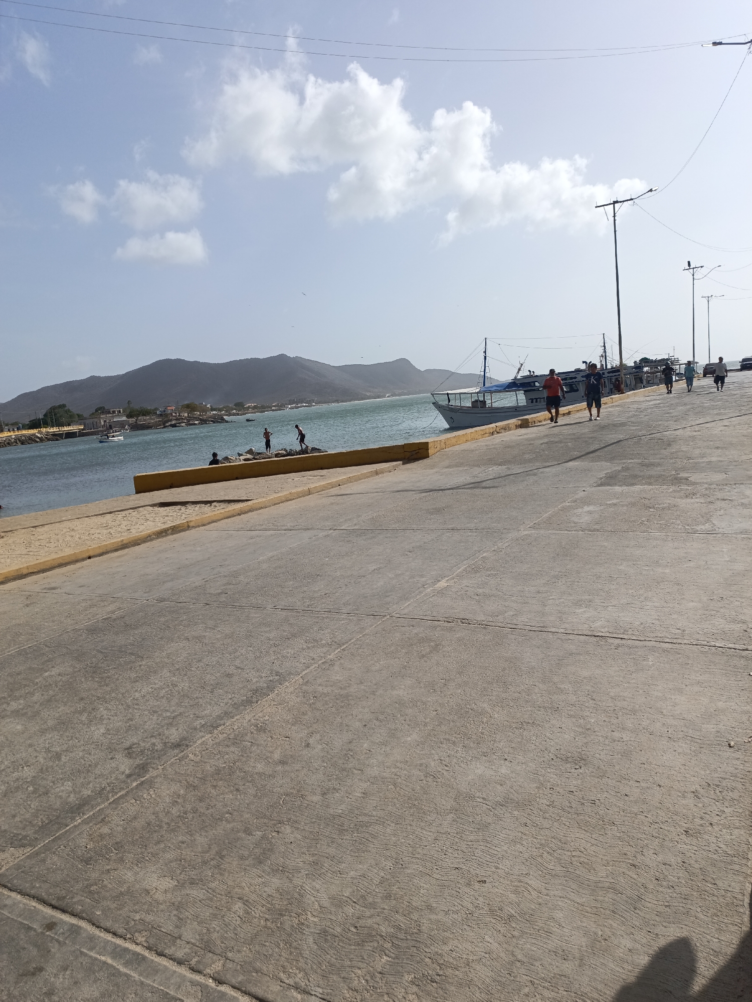 The image depicts a paved area in Juan Griego, Venezuela, possibly a dock or promenade, with a slight incline. The foreground is dominated by the pavement, marked with seams. A yellow curb defines the edge, separating the pavement from the water. In the midground, several people are walking along the paved area, while others are closer to the water, interacting with rocks. A boat is docked nearby. The background includes a body of water, a stretch of land with buildings, and a mountain range. The sky is partially cloudy with light blue patches. Utility poles and wires are also visible.