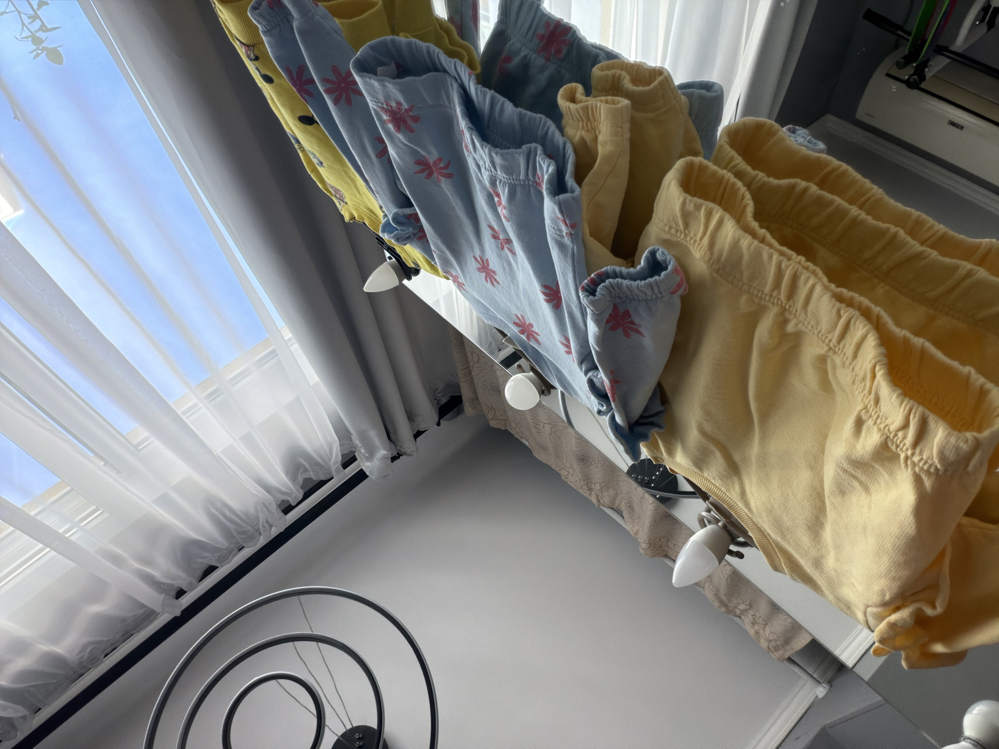 The image captures an indoor scene from a low, tilted angle, looking upwards towards a brightly lit window. The primary subjects are several pieces of children's clothing, possibly pants or shorts, hanging on a white rack positioned beneath the window. Some garments are plain light yellow, while others are light blue with recurring pink floral or star-like patterns. These clothes appear to be air-drying or neatly stored.
Through the window, a clear, vivid blue sky is visible, suggesting it is daytime with good weather outside. The window is adorned with translucent white curtains or blinds, allowing natural light to flood the space. In the lower part of the frame, a light-colored floor is visible, along with a decorative metal object featuring concentric circles or a spiral design, possibly part of a light fixture or a fan. To the far right, partially obscured by the hanging clothes, an air conditioning unit can be seen mounted on a wall. The overall setting is an interior space, likely a home, potentially in a child's room or a utility area, hinting at a modern dwelling in a location like Sochi, Russia, which benefits from natural light. No people or visible text are present in the image.