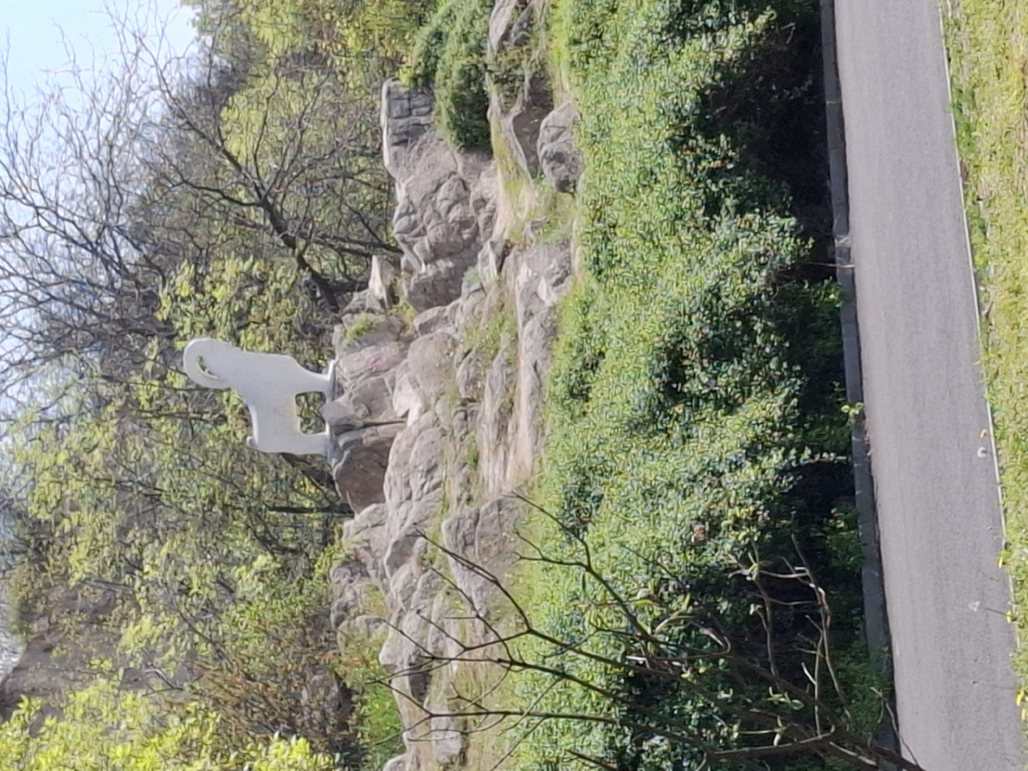 The image shows a white sculpture of a horse on a rocky outcrop in Plovdiv, Bulgaria. The sculpture is positioned on a rock formation covered with green foliage. To the right of the rocks is a paved pathway. Bare trees and other greenery fill the left side of the frame. The sky is light blue and visible in the upper left corner.
