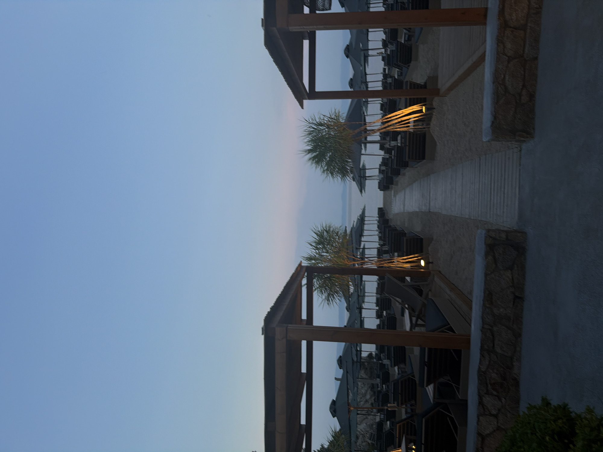 The image depicts a beach area in the Sithonia Municipal Unit, Greece, at dusk or early dawn. The sky is a clear, soft blue, transitioning to a lighter hue near the horizon where it meets the sea. In the foreground, a sandy beach is lined with numerous empty sun loungers and dark umbrellas, arranged in neat rows under thatched-roof cabanas. A wooden boardwalk leads towards the sea, separating the seating areas. Two prominent wooden cabanas, one on the left and one on the right, frame the view. Palm trees with sparse fronds are visible between the sunbeds, and small, warm-toned lights illuminate the scene. A low stone wall runs along the bottom of the frame, separating the beach from a paved pathway. No people are present in the image. The overall atmosphere is serene and tranquil, suggesting a quiet, unoccupied beach resort.