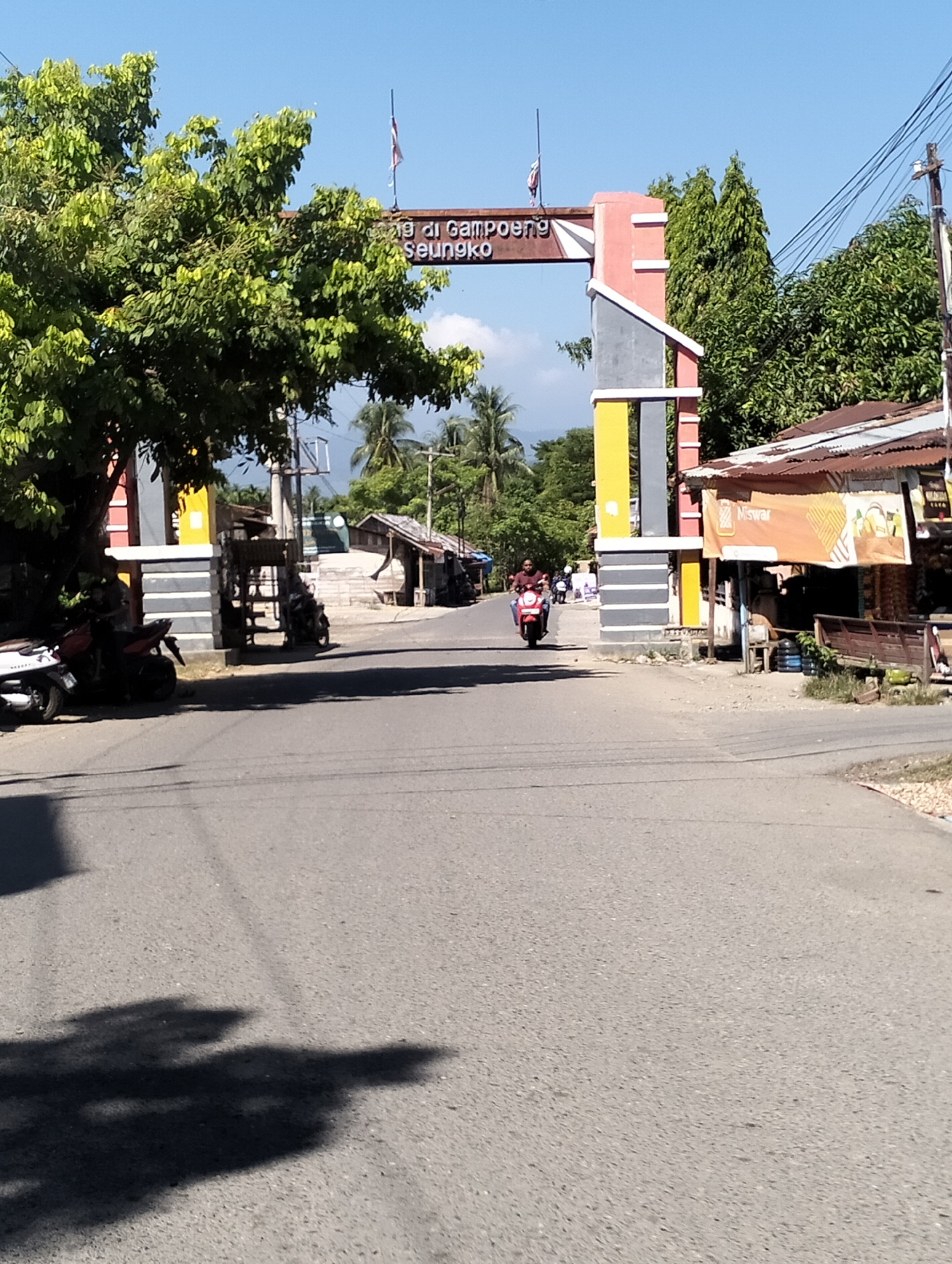 This image depicts an outdoor scene on a clear, sunny day, likely in Keudejunieb, Indonesia, suggested by the presence of an archway with Indonesian text. The archway, constructed with red, gray, and yellow elements, spans across a paved road. The text on the archway reads "Gampong Seunöngko," indicating the name of a village or settlement.
In the foreground, a portion of the road is visible, with shadows from trees cast upon it. To the left of the road, a motorcycle is parked near a large tree with lush green foliage. Further down the road, a person is riding a red scooter, approaching the archway.
To the right of the road, a building with a corrugated metal roof is visible, with an orange banner displaying the word "Miswar" in prominent lettering. Several smaller structures and trees are present in the background. Utility poles and overhead wires are also visible, indicating the presence of infrastructure. The overall environment appears to be a semi-rural or suburban area.