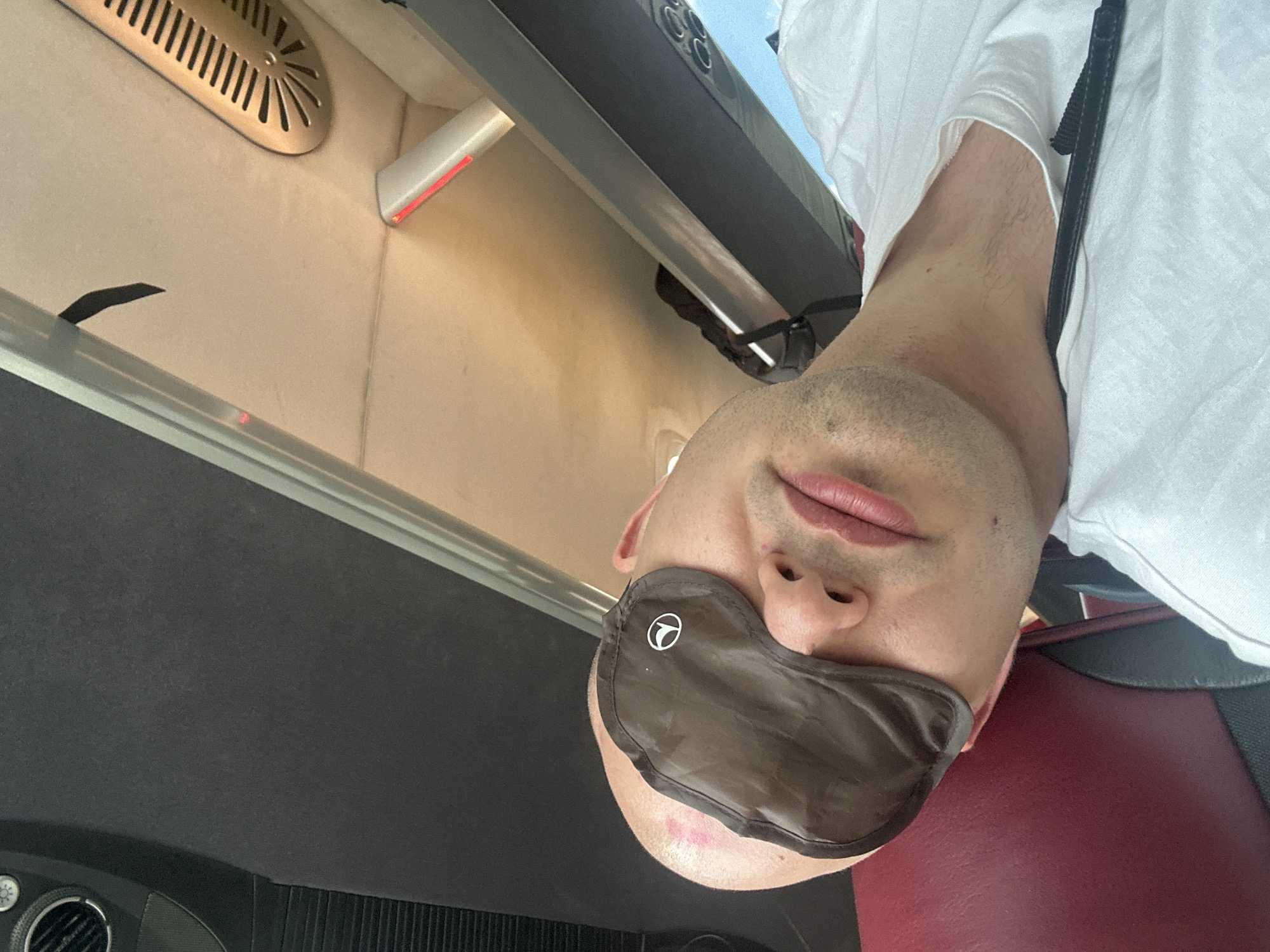 A close-up, overhead, low-angle shot shows a man wearing a brown sleeping mask over his eyes. He is lying down, his head tilted to the left. His skin is pale, and he has stubble on his chin and jawline. His lips are parted. He is wearing a white t-shirt. The background appears to be the interior of a vehicle, possibly a bus or van, with dark grey upholstery above him, and a red seat cushion beneath him. A silver-colored bar runs diagonally across the frame, and a metallic vent with a decorative pattern is visible on the ceiling. The lighting suggests an indoor environment, possibly during daytime, with some light sources casting a warm glow. There is no discernible indication of the city of Arnavutköy, Turkey.