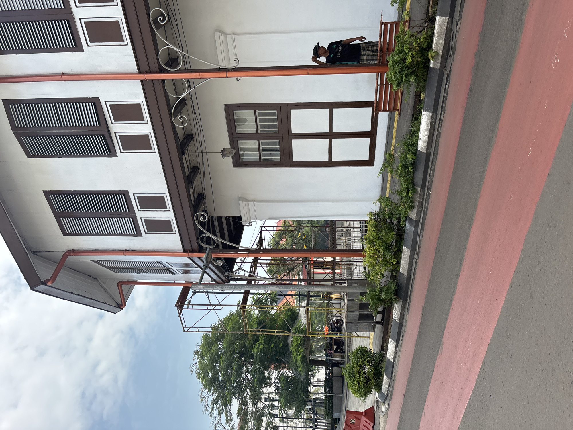 An outdoor urban scene in West Jakarta, Indonesia, captures a street view dominated by a two-story building on the right. The building features a white facade with dark brown wooden window frames and louvered shutters on the upper floor, interspersed with smaller rectangular decorative panels. A prominent orange drainage pipe runs horizontally across the upper part of the building, attached to a white decorative metal bracket. Below this, a large brown-framed window with multiple panes is visible on the first floor.
A person, likely an adult male, stands near the building's entrance or a recessed area on the first floor. He is wearing a black t-shirt with "EMIRATES" visible across the chest, plaid shorts, and a dark cap. He is leaning against a low reddish-brown railing or wall section.
To the left of the building is a paved street featuring broad, alternating red and dark gray stripes, indicative of a pedestrian crossing or specific road marking. A curb separates the street from the building, marked with a distinctive black and white pattern. Small green bushes and plants are neatly arranged along this curb. In the background, beyond the street, scaffolding is visible, suggesting ongoing construction or maintenance. Large green trees and glimpses of other buildings complete the distant view under a bright, partly cloudy sky, indicating daytime.