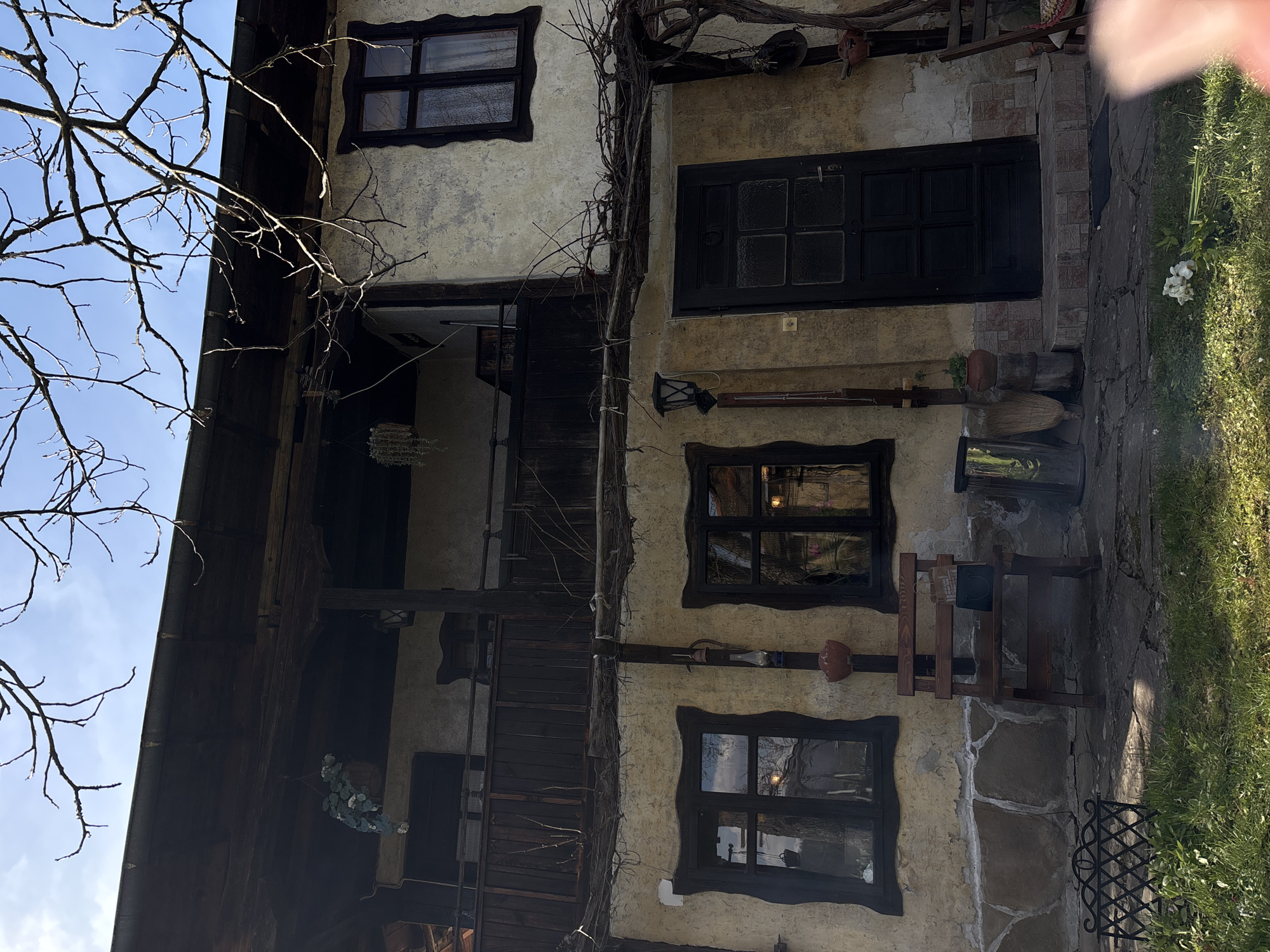 Exterior shot of a building in Gorno Trape, Bulgaria, featuring a three-story structure with a facade displaying textured plaster and dark wooden window frames. There are at least three visible windows, each with a distinct dark outline. A dark-colored front door is centrally located on the second level. A dried vine climbs the front of the building. The architecture includes a balcony or elevated walkway on the second story. Bare tree branches are visible in the upper left. A patch of grass and a stone-paved pathway can be seen to the right. Various decorative items are mounted on the walls and along the facade.