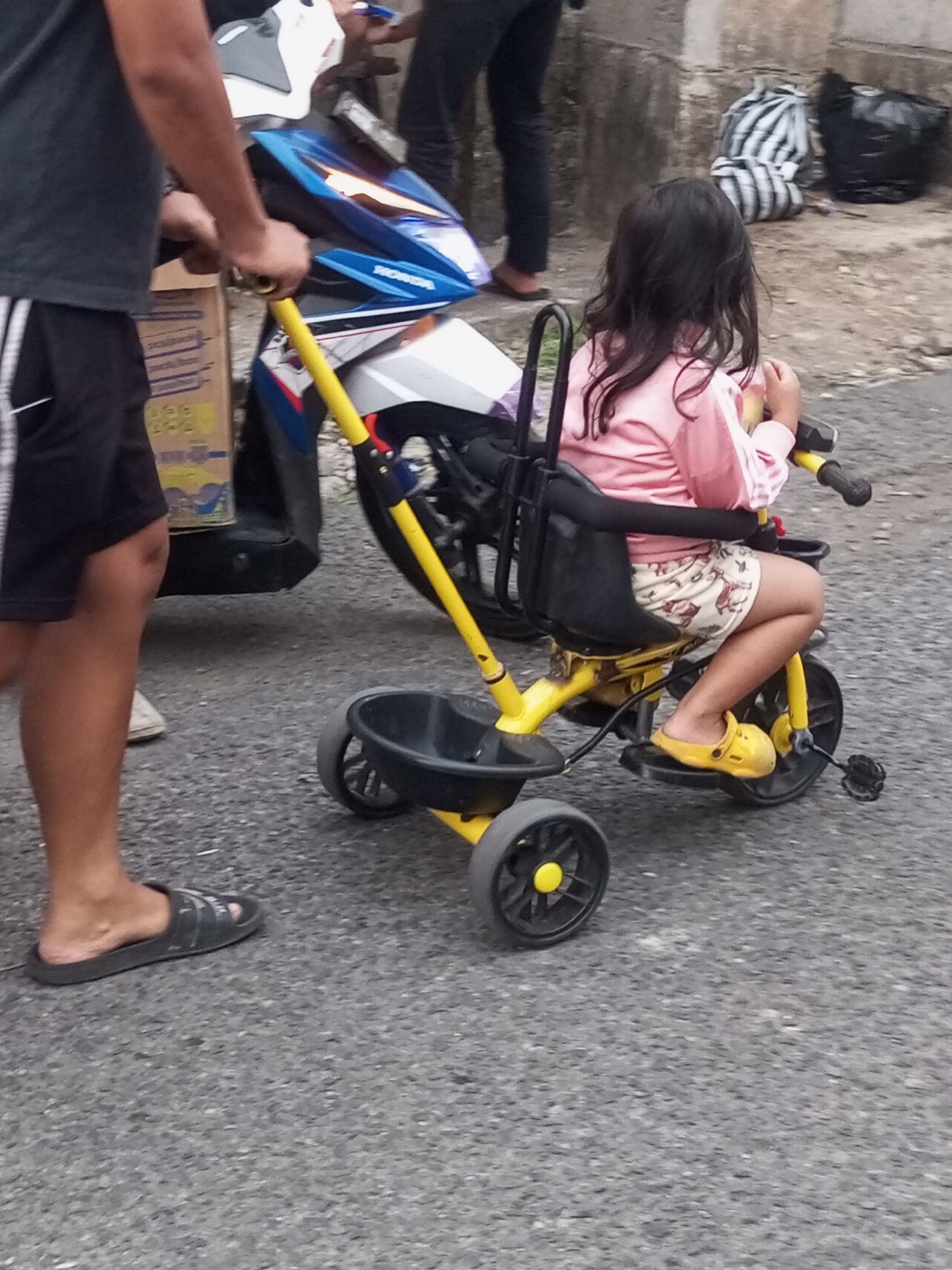 In Pandansari, Indonesia, an outdoor scene depicts a young girl seated in a yellow child's tricycle. The tricycle has a black seat and a black basket at the front. The girl wears a pink long-sleeved shirt and patterned shorts. Her feet are clad in yellow clogs. To the left of the tricycle, a person's legs are visible. This individual is wearing black shorts with white stripes and dark gray flip-flops. They are holding onto a yellow bar that appears to be part of a scooter or motorcycle, which is partially visible behind them and to the left. The scooter is blue and white. The ground is asphalt. In the background, several large, dark-colored trash bags are visible on the ground, along with a patterned bag that looks like it contains clothes. The wall behind the trash bags is made of rough, grey stone.
