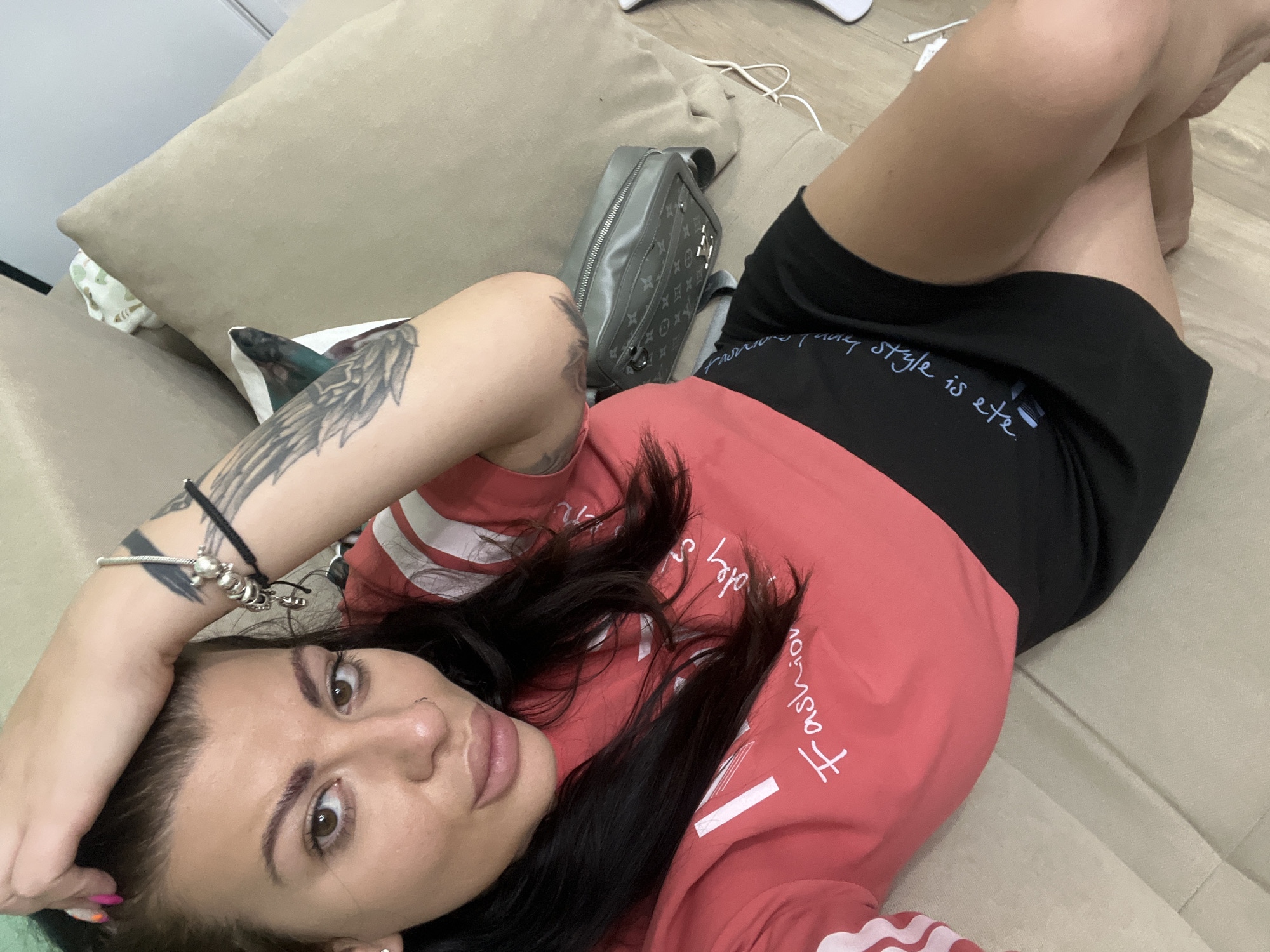 A woman is lying on a beige sofa in a room. She is wearing a coral t-shirt with white stripes on the sleeves and a black skirt. She has dark hair, brown eyes, and is wearing a nose ring and several bracelets on her left wrist. Her left arm is resting on her head. A grey bag with a patterned design is on the sofa next to her. The room appears to be a living space. There is no indication of the city of Plovdiv, Bulgaria in the image.
