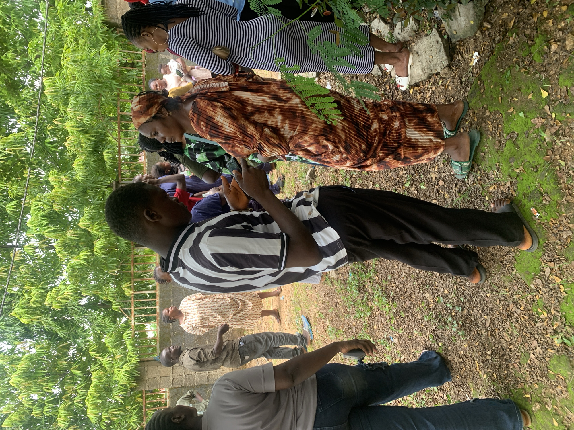 In Akwanga, Nigeria, a group of people are gathered outdoors on a dirt ground covered with leaves and sparse green vegetation. A man wearing a black and white striped shirt and black pants is holding a mobile phone, seemingly engaged in a conversation with a woman. The woman is wearing a brown and white patterned dress and a headscarf. To her right, a woman in a black and white striped dress stands, and to the left, a person in a gray t-shirt and blue jeans faces away from the camera. Other individuals are visible in the background, with a fence and lush green foliage forming the backdrop. The lighting suggests it is daytime.