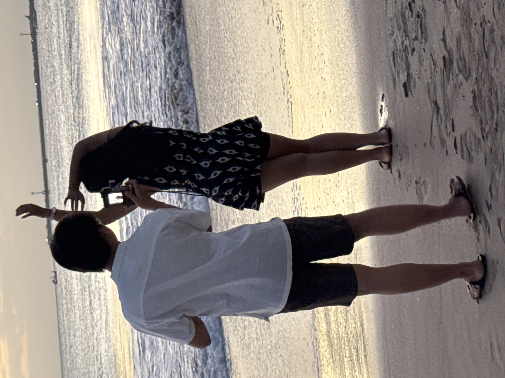 Two individuals are captured from behind on a sandy beach, with the ocean and the sky in the background. The person on the left, wearing a white short-sleeved shirt and dark shorts, stands with their back to the camera, facing the water. Their left arm is slightly raised. The person on the right, dressed in a dark-patterned dress, is holding a camera and appears to be taking a photograph. Their right arm is raised above their head, and their left arm is bent as they hold the camera. The water shows gentle waves and the sky is a pale yellow, suggesting either sunrise or sunset. The sandy beach in the foreground is wet in places and shows patterns of sand and water. The overall scene conveys a relaxed, vacation-like atmosphere. This scene is likely taking place in Kuta, Bali, Indonesia, a well-known beach destination.