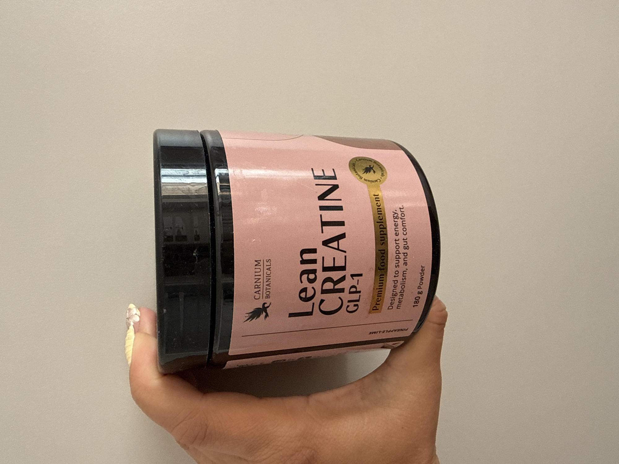 A hand holds a pink and black tub of Carnium Botanicals Lean Creatine GLP-1, a premium food supplement. The tub has a black lid and a pink label with black text. Text on the label includes "Carnium Botanicals," "Lean CREATINE," "GLP-1," "Premium food supplement," "Designed to support energy, metabolism, and gut comfort," and "180 g Powder." There is also a small circular seal with an image of a bird and text that reads "Carnium Botanicals." A small portion of the thumb and index finger are visible, with some fingernails at the top of the frame. The background is a plain, light-colored wall.