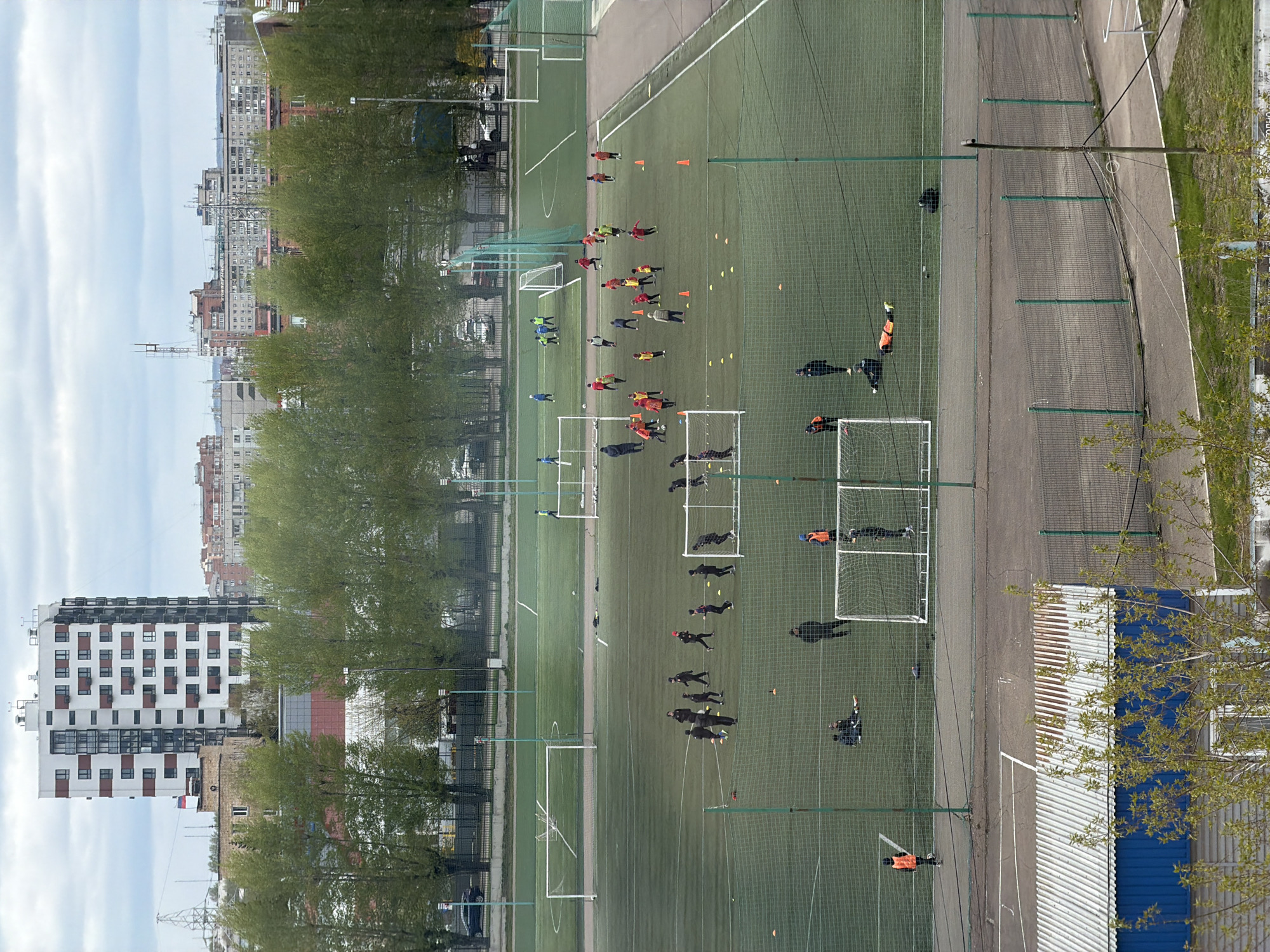 The image shows a soccer field in Krasnoyarsk, Russia. The field has multiple goals set up, with groups of people of varying ages training in different sections. Some are in groups, while others practice individually. Cones are scattered around as training aids. A paved walkway runs along one side of the field, separated by a net fence. The buildings of Krasnoyarsk line the left side of the image, with trees dividing the field from the city. A building with a blue and white corrugated metal roof and emerging tree branches is on the right.