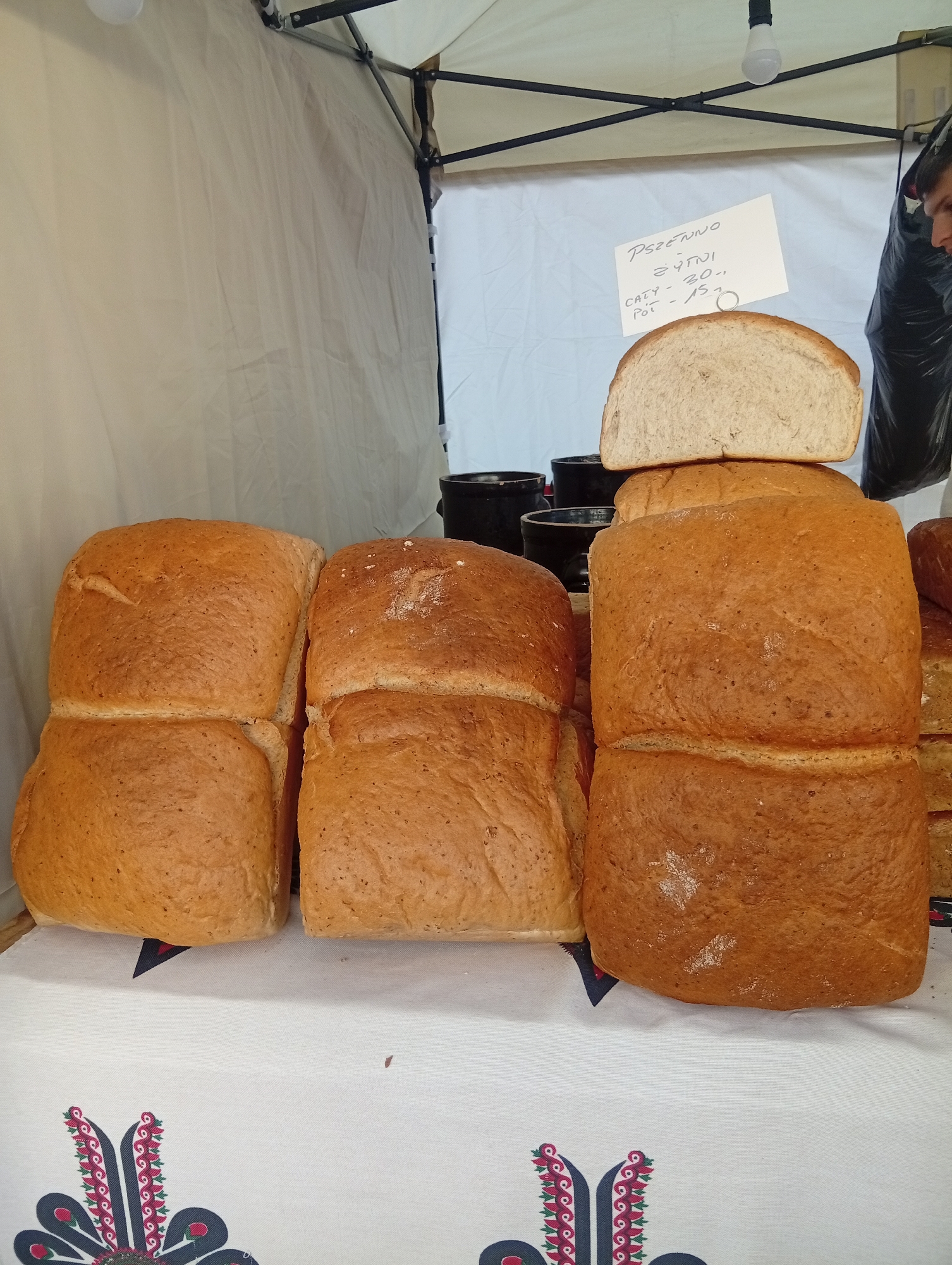 A close-up, eye-level shot shows a stack of loaves of bread on display at an outdoor market stall, likely in Wrocław, Poland, based on the Polish writing on the sign. The breads are golden brown and appear to be whole grain. In the foreground, four loaves are stacked in two columns. A slice of bread is visible on top of the right column. Behind the loaves, there are black containers and a white tarp that forms the backdrop of the stall. A sign, handwritten in Polish, is attached to a metal pole above the bread. The sign reads "PSZETNWO ŻYTNI CAŁY - 30,- PÓŁ - 15,-". A person is partially visible on the right side of the image, wearing a black trash bag over their head and shoulders, suggesting they might be working at the stall. The table is covered with a white cloth featuring a colorful folk art pattern.