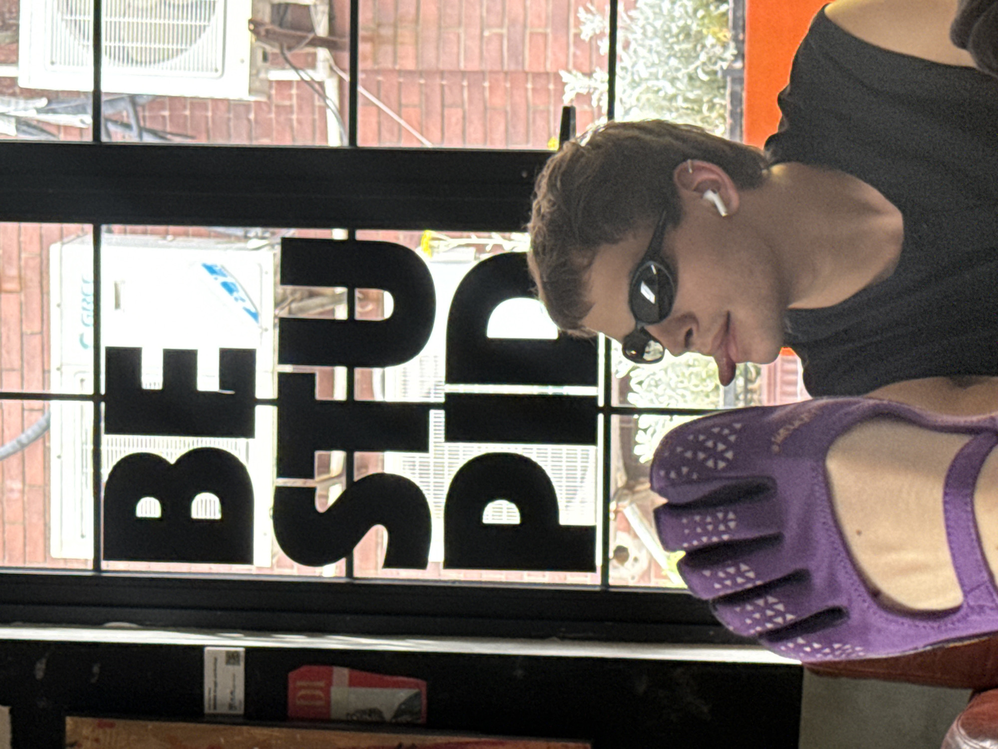 The image, rotated 90 degrees counter-clockwise, captures a person looking out through a window adorned with large, black letters. The person, appearing to be a young male, has short dark hair, wears dark sunglasses, and a black t-shirt. A white earbud is visible in his right ear. His head is tilted downwards, suggesting a relaxed posture or observation of something outside the frame.
In the immediate foreground, a vibrant purple "toe shoe" (minimalist footwear) with white geometric patterns is prominently displayed, likely belonging to the same individual.
The window features bold black cut-out letters spelling "BEST PIZZA". Beyond the window, an outdoor urban scene unfolds, revealing a brick building wall, two white air conditioning units (one partially branded "GREE"), and some green foliage, consistent with a setting in Putuo District, China. The scene is brightly lit by natural daylight. Below the window, a dark surface, possibly a shelf or counter, holds a small cardboard box. The overall impression is a casual, daytime setting, possibly inside a cafe or restaurant.