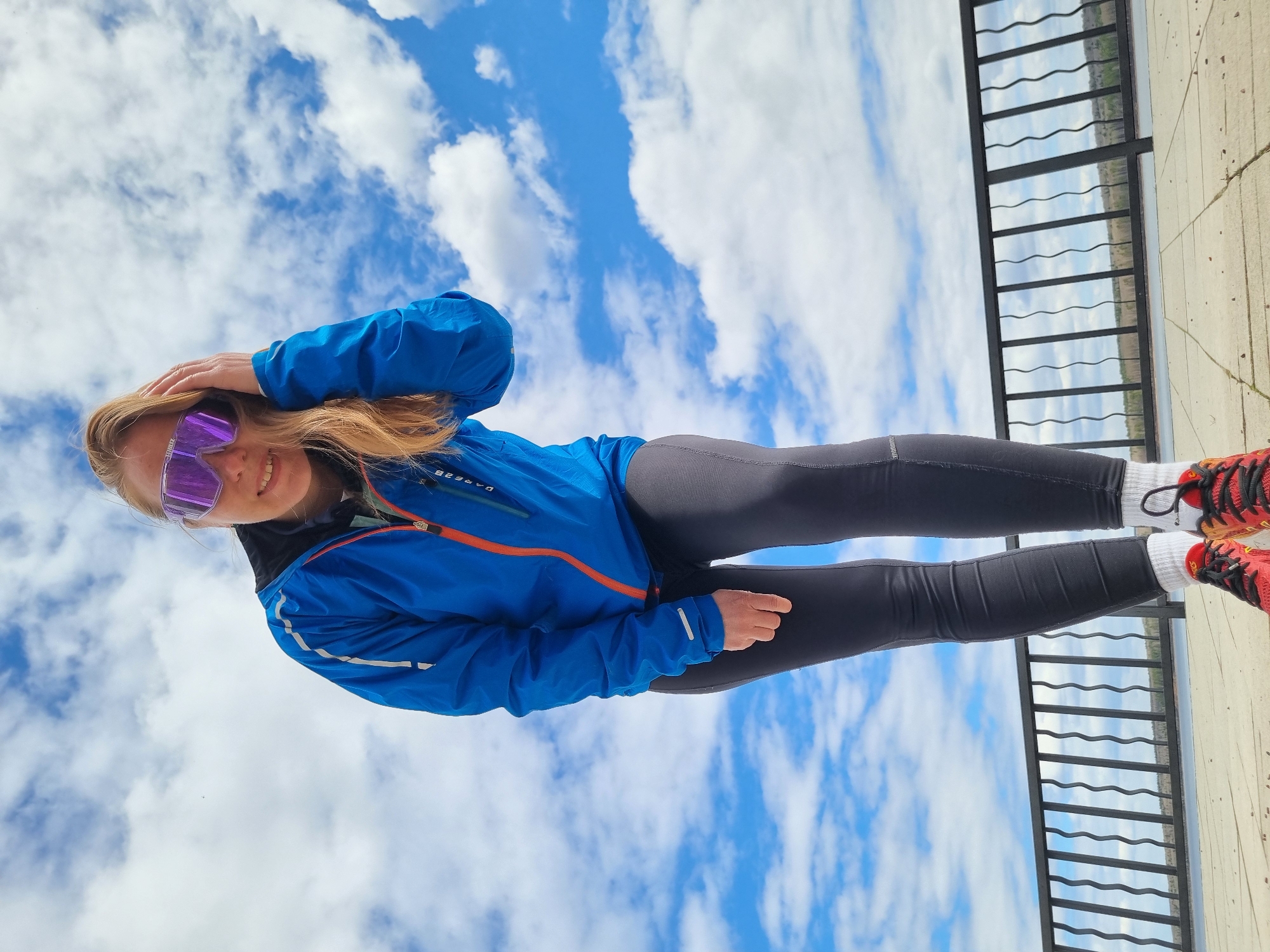 A woman wearing a blue jacket, black leggings, white socks, and red and orange running shoes stands on a light-colored concrete surface with a black metal railing in the background. The background beyond the railing shows a line of trees, likely indicating a park or natural area. The sky is visible in the background, filled with white and blue clouds. The woman is wearing reflective purple sunglasses and has her hand on her head, appearing to adjust her hair or sunglasses. The angle of the image suggests it was taken from a low perspective, looking upward. Given the location of Chaikovsky, Russia, the scene likely depicts an urban environment near a park or natural area on a day with partly cloudy skies.