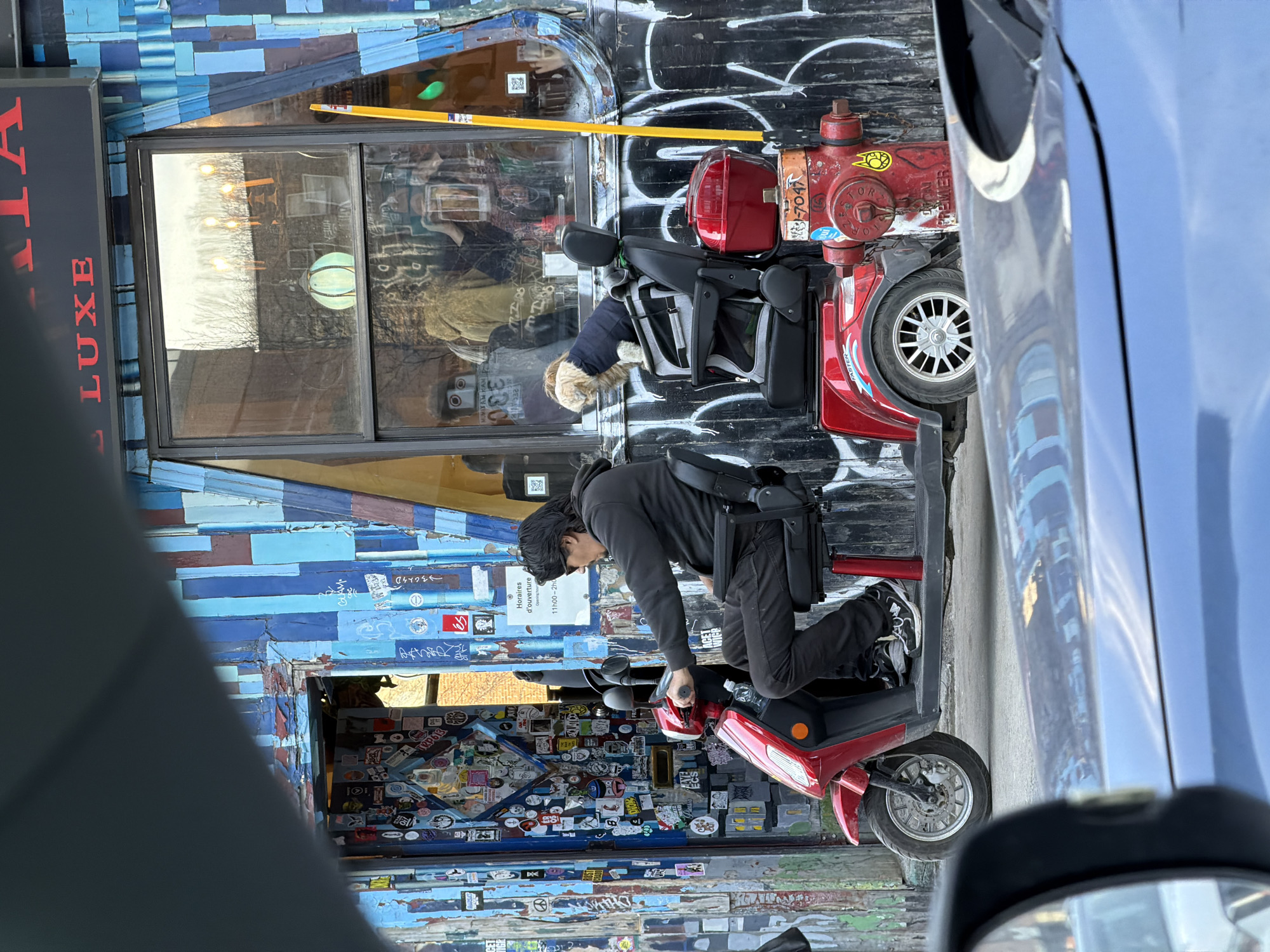 Here's a detailed description of the image:
**Scene:** The image appears to be taken from inside a vehicle, likely a car, looking out at a street scene. The primary focus is a person on a red mobility scooter parked beside a building with distinctive features.
**Location:** Based on the signage and architectural style, the location is likely in Montreal, Canada.
**People:** There is a person, presumably an adult, dressed in dark clothing (hoodie and pants) operating the red mobility scooter. Their face is partially obscured, and they are bent over the scooter's handlebars. A dog is sitting on a mobility scooter as well. There is a reflection in the window that may be a person.
**Subjects and Objects:**
* Red mobility scooter: It's parked on the street next to the building.
* Building: The building has an eclectic facade. The lower portion of the building's exterior is covered in numerous stickers, while the upper portion features a collage of painted blue and brown wooden planks. The building also has a window with signage visible inside.
* Fire hydrant: A red fire hydrant is located next to the building.
* Car: A portion of a blue car is visible on the right side of the frame, partially obscuring the scene.
* Street: The street surface appears paved.
**Actions and Interactions:** The person is interacting with the mobility scooter, potentially starting it or adjusting controls.
**Environment:** The scene is an urban environment, with the building and vehicle suggesting a street setting. The presence of the scooter and hydrant indicates a street-side location. The lighting suggests it is daytime.
**Signage/Text:** The building features signage that says "ACET...LUXE". There are stickers on the door, which includes one that says "Faster" in the image. A yellow measure is placed next to the window.