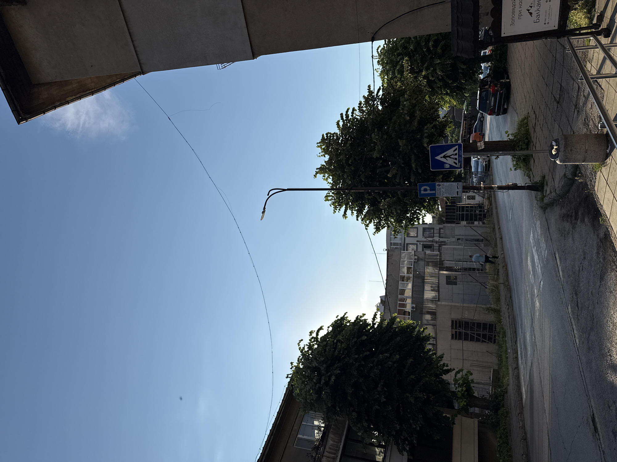 The image depicts a street scene in Vratsa, Bulgaria. On the right side of the image, a concrete building with a dark roof is visible, with a white sign mounted on a black frame. The sign contains text in Cyrillic, and some of the text appears to read "Балкан" and a phone number. In the foreground, a sidewalk with a metal railing is present, with a cylindrical concrete trash receptacle. Further down the street, a pedestrian crossing is marked on the asphalt road. A street lamp with two lights stands on the left side of the road, next to a blue parking sign with Cyrillic text and a white square sign with a pedestrian symbol. Several trees with dense green foliage line the sidewalk and extend into the background. On the left side of the image, a building with multiple windows and a balcony is visible. In the middle ground, there is a construction site with scaffolding and concrete structures. A person in a light blue shirt and dark pants is walking on the sidewalk towards the construction site. Several cars are parked on the street and on the sidewalk. The sky above is clear blue with a few wispy clouds.