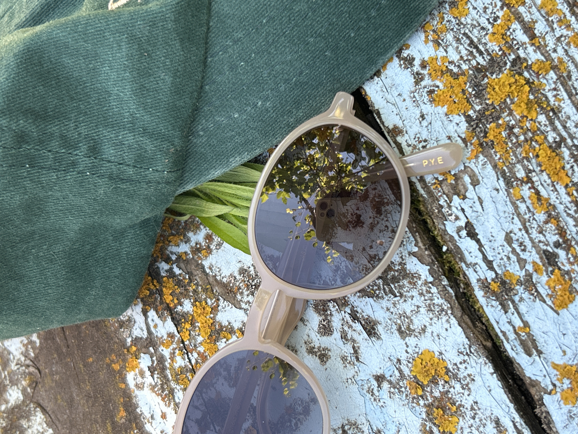 The image features a pair of light-colored sunglasses resting on a weathered wooden surface. The wood is painted a pale blue, with patches of peeling paint and textured yellow lichen, suggesting an outdoor setting. To the left of the sunglasses, a portion of a dark green cloth, possibly a hat or bag, is visible. Green foliage peeks out from beneath the cloth and the sunglasses. The lenses of the sunglasses reflect the surroundings, showing a clear sky and the branches of trees. On one of the sunglasses' arms, the text "P.Y.E" is visible in gold lettering. The lighting suggests it is daytime, with sunlight illuminating the scene and creating reflections. There is no discernible activity taking place other than the objects being placed on the surface. The overall impression is one of casual repose in an outdoor environment, possibly during warm weather. The location context of Zvonarevka, Russia, does not provide specific visual cues within the image itself.
