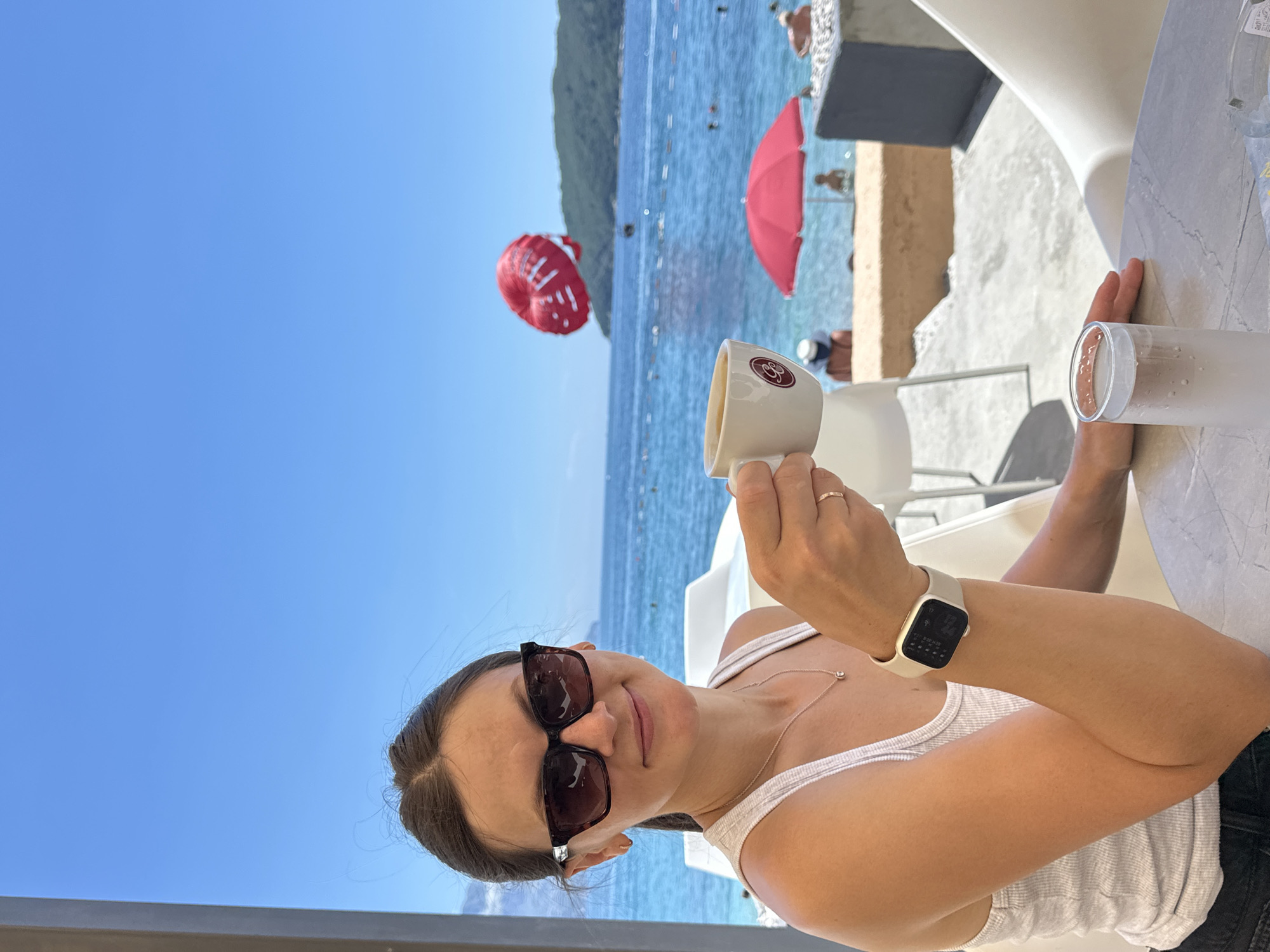 A woman with brown hair pulled back and wearing sunglasses, a white tank top, and a necklace sits at a table with a cup of coffee and a glass of water. The table is outdoors, likely at a cafe or restaurant with a view of the sea. In the background, the ocean is visible with several people in the water, a red umbrella, and a red parasailer in the sky. The scene appears to be Budva, Montenegro, given the typical coastal scenery and activities associated with the location.