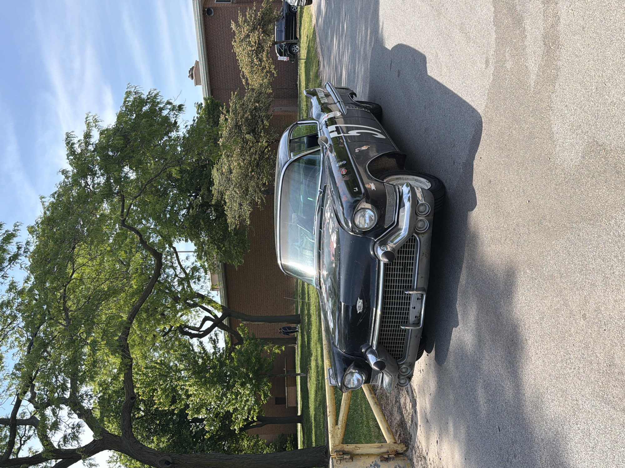 The image depicts a vintage black Cadillac parked on an asphalt road in a park-like setting. A large, leafy tree partially obscures the view of a brick building in the background. A yellow barrier is in front of the car. The Cadillac features chrome detailing on its front grill and headlights, with stickers adorning its body. There is another black vehicle visible in the background. The location appears to be Chicago, United States of America based on the style of the buildings, vehicles, and the general urban park environment, but there is no specific landmark that confirms this information in the image.