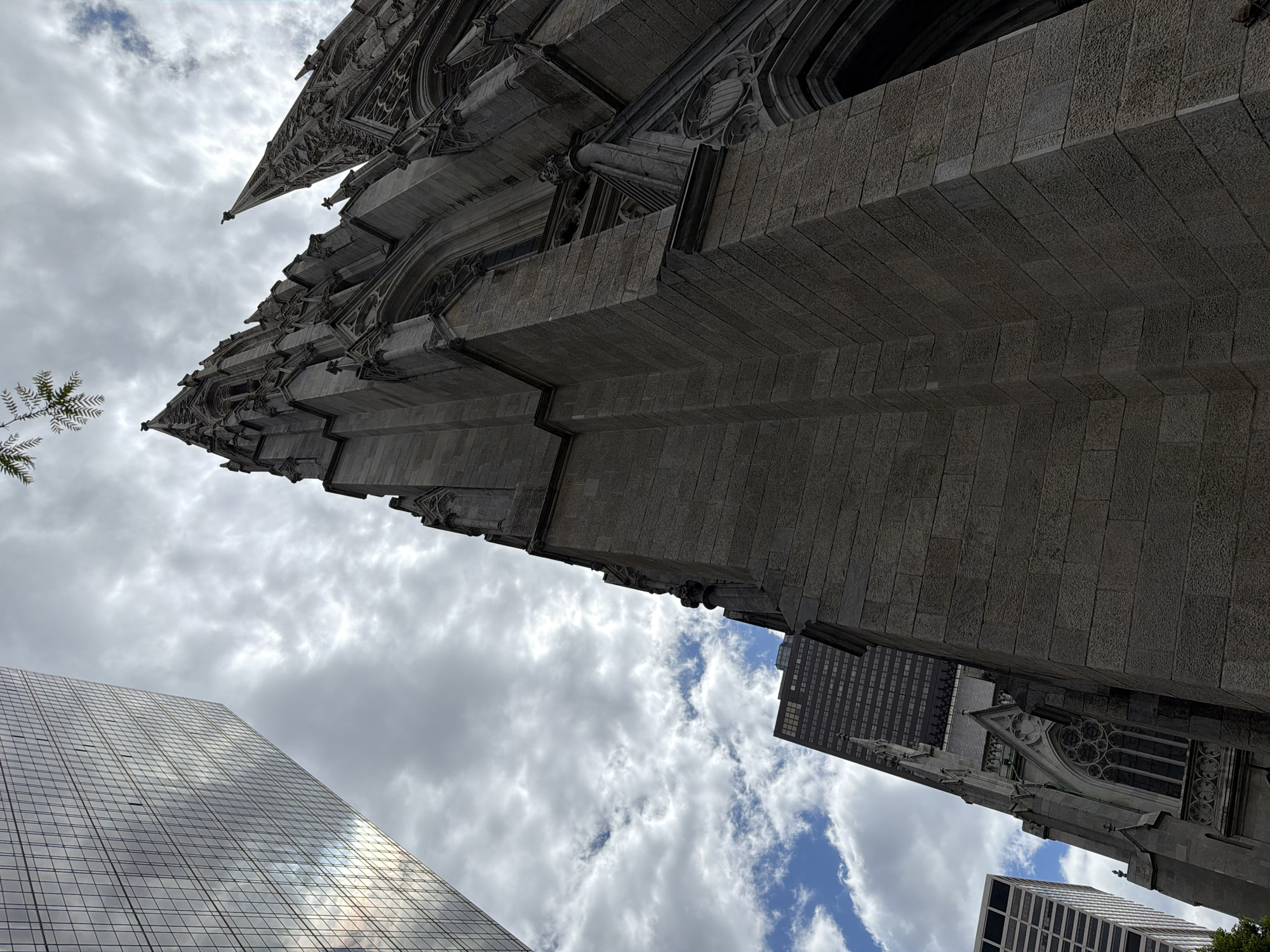 The photograph captures a low-angle view of a Gothic-style church, possibly St. Patrick's Cathedral, situated in New York City, United States of America. The stone facade of the church, characterized by its vertical emphasis and architectural details, dominates the frame. The sky is filled with clouds, and portions of nearby modern skyscrapers are visible, contrasting with the traditional architecture of the church. A few tree leaves are seen at the left, partially in view. The composition suggests an attempt to juxtapose historical and contemporary elements within the urban landscape.