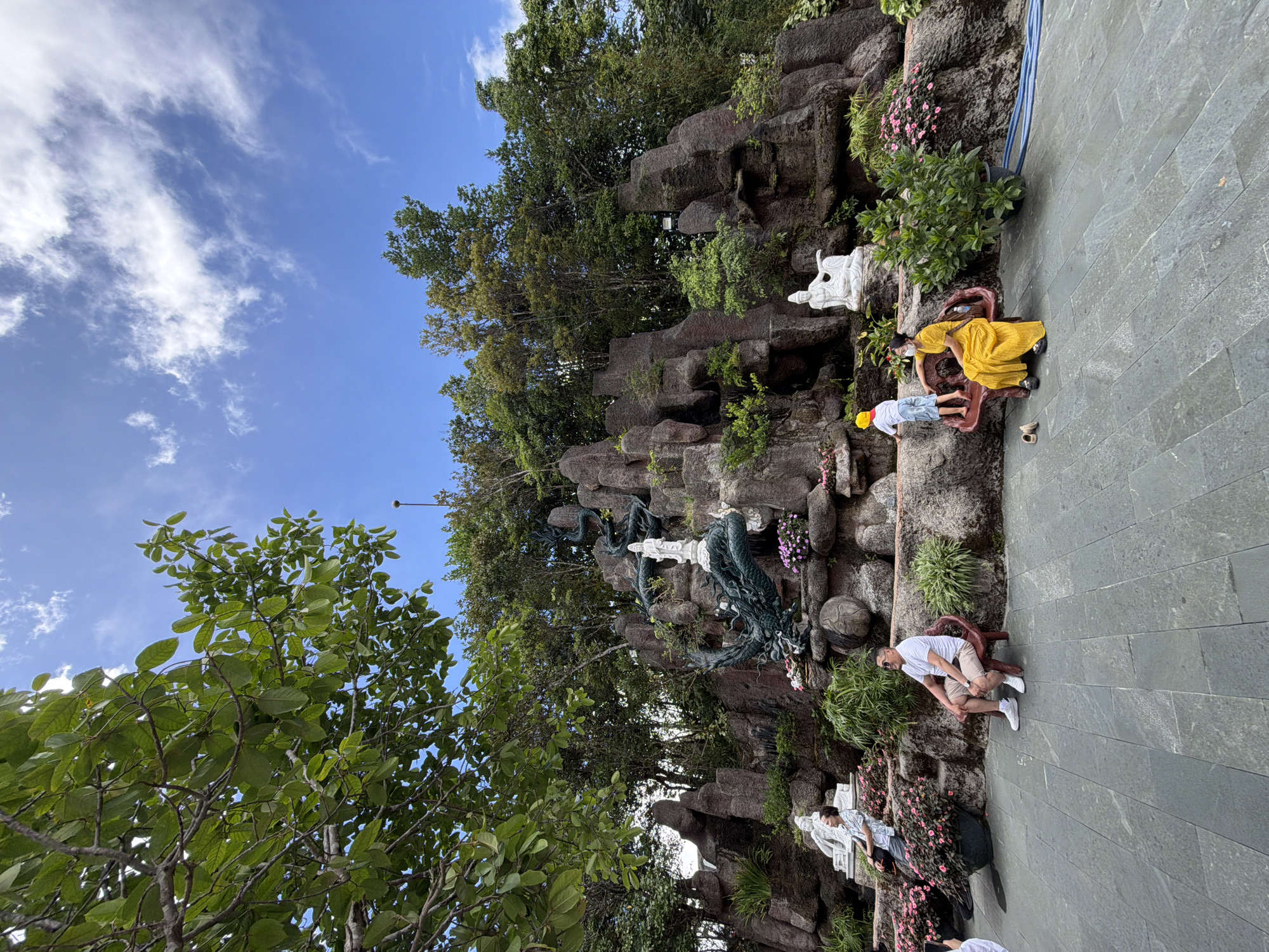 This image captures a scene at the Dragon Bridge's vicinity in Da Nang, Vietnam, depicting a man and a woman sitting on benches. The man, wearing a white t-shirt and beige shorts, is seated on a red bench. The woman, dressed in a bright yellow dress and a white face mask, is also on a bench, with a young child in a white shirt and blue shorts standing near her. Behind them, a large rock formation is adorned with a statue of a dragon and several Buddhist figures, suggesting it is part of a temple or cultural attraction. Lush green trees and vibrant pink flowers surround the area, and the sky above is a clear blue with scattered white clouds. The ground is paved with large grey tiles, and a blue hose is visible on the right side of the frame.
