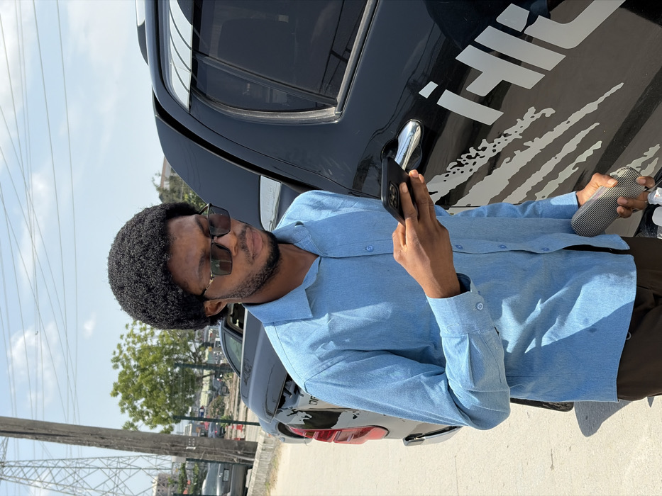 In the city of Igboefon, Nigeria, a young adult male stands outdoors, facing slightly left of center, observed from the chest up. He has a dark afro hairstyle, a slight beard, and wears dark rectangular sunglasses with gold-colored accents, a light blue long-sleeved button-up shirt, and dark trousers. He holds a black smartphone horizontally in his left hand, actively looking down at its screen. His right hand holds a striped, dark-colored case and a small white object. He is positioned next to the driver's side of a dark-colored pickup truck or SUV, which features a chrome door handle and a white "claw mark" style decal with partial white lettering ("i-Hili") on its side. The background reveals an urban environment under a clear sky with scattered clouds. A large metal utility pole with overhead lines is visible on the far left. Additional vehicles and green foliage are present further in the background, suggesting a roadside or parking area. The scene is brightly lit by natural sunlight, casting discernible shadows.
