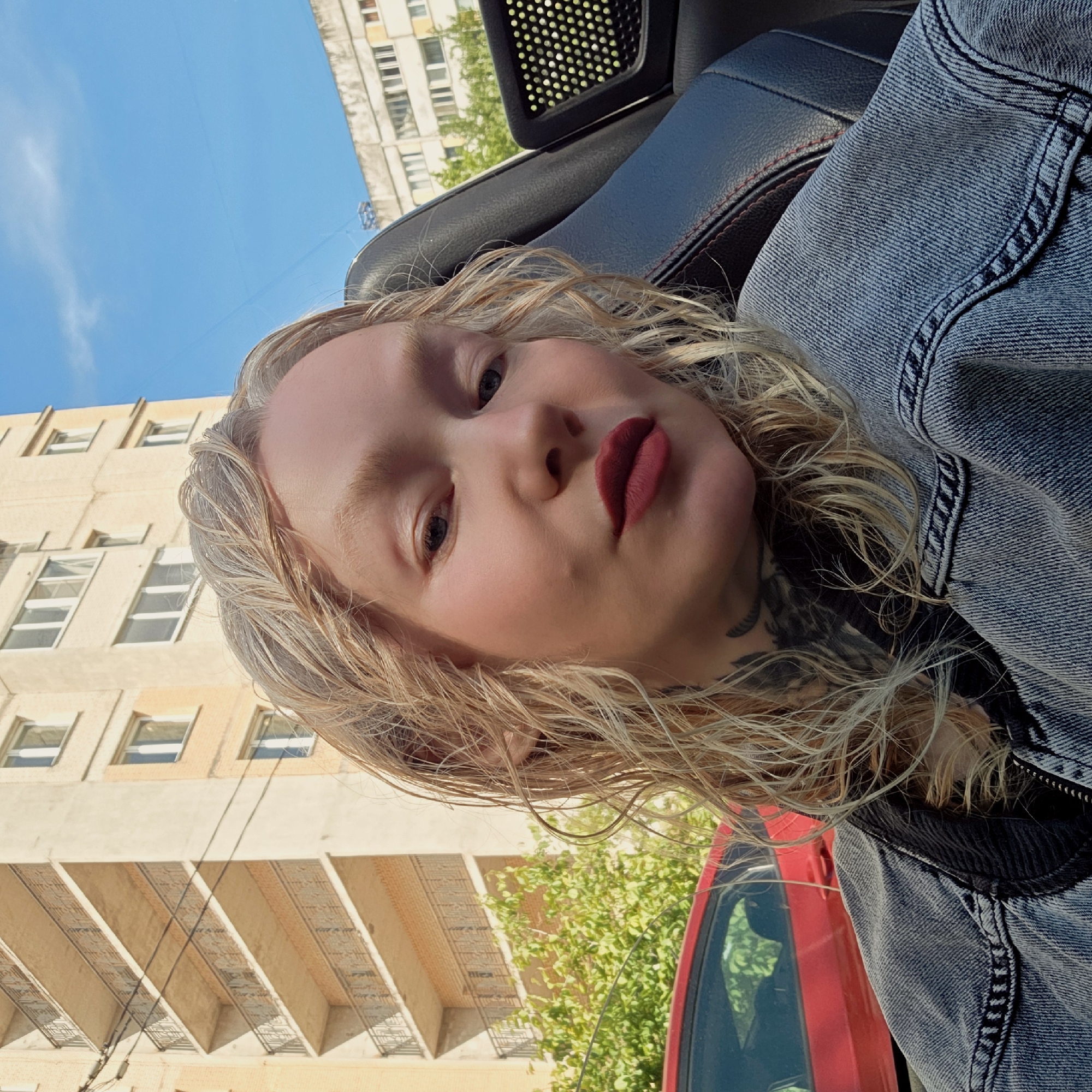 A fair-skinned person with blonde, wavy hair and dark red lipstick is seated in a car. They are wearing a blue denim jacket. Tattoos are visible on their neck. The car's interior is black with red stitching on the seats. Outside the car, there is a multi-story building with numerous windows, and a blue sky is visible. The location is Saint Petersburg, Russia.