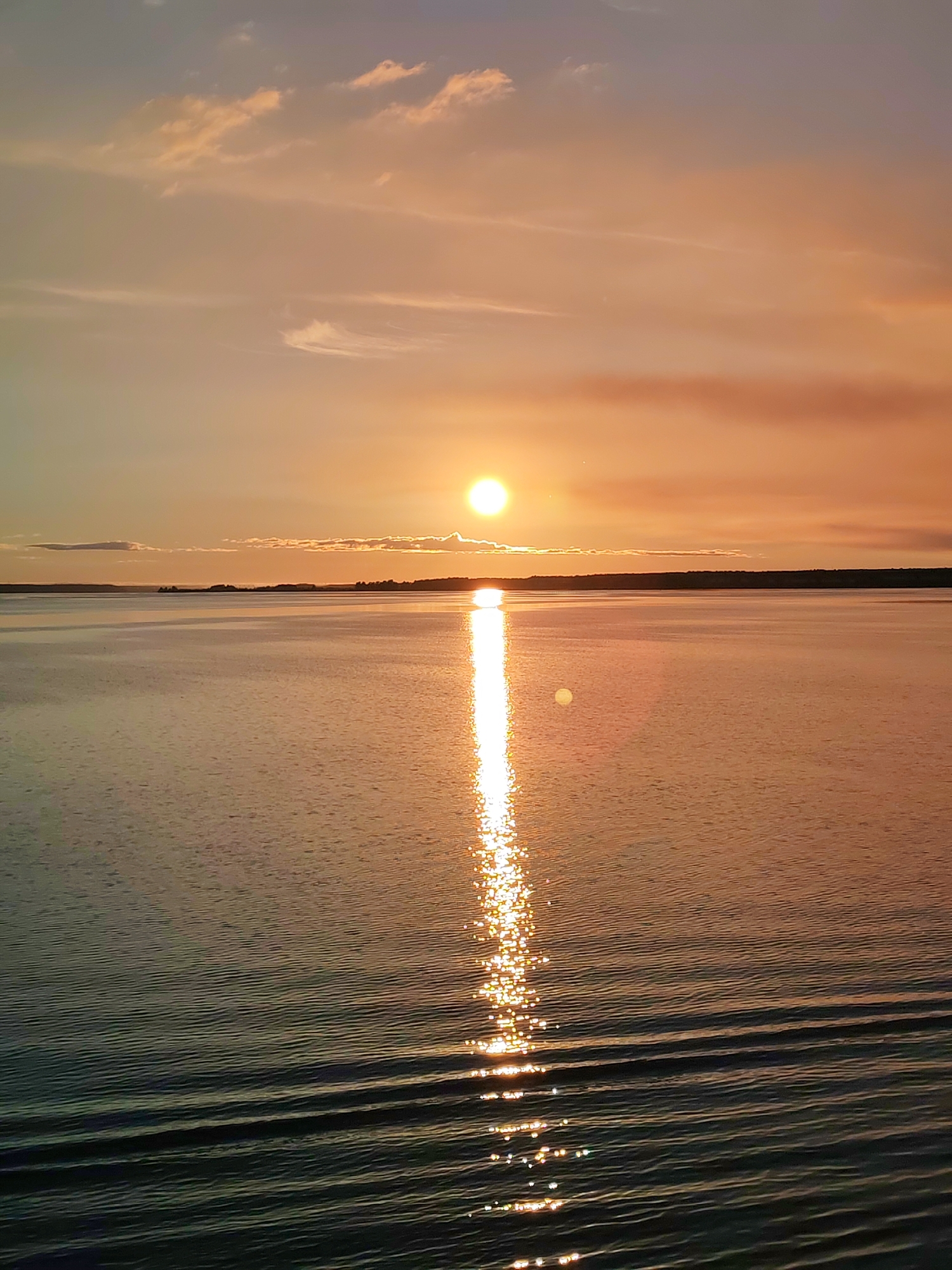 The image shows a sunset over a body of water. The sun is positioned near the horizon, reflecting a bright path of light across the water's surface. The sky is a mix of orange, pink, and light blue hues, with some scattered clouds. The water has gentle ripples. A dark line of land is visible in the distance.