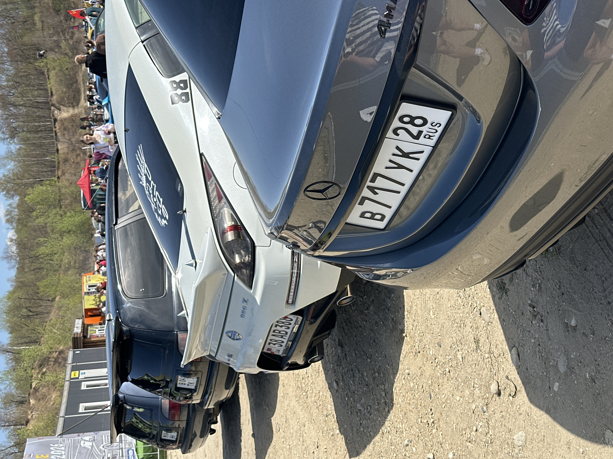 The image shows a row of parked cars, including a grey Mercedes-Benz with the license plate "B717YK28" (RUS) and a white Nissan 350Z with the license plate "38 AB 388". A crowd of people is visible in the background. The location appears to be outdoors, with a dirt or gravel surface. Buildings are visible in the background. Based on the license plate information, it is inferred that this scene could be in Chigiri, Russia.