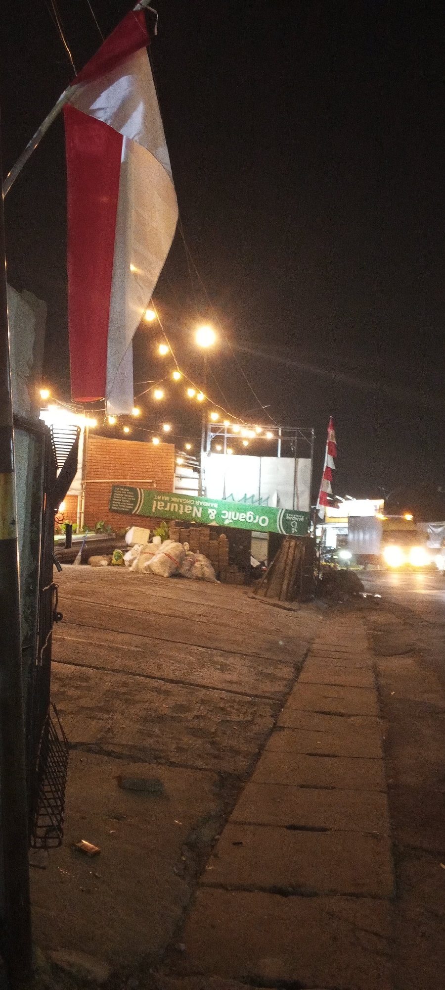 This is a photograph taken at night of a street scene, likely in Depok, Indonesia. In the foreground, a red and white flag is prominently displayed, hanging from a pole. The flag appears to be the Indonesian flag. String lights are strung across the scene, casting a warm glow. A sign in green lettering reads "Organic & Natural BANDAR ORGANIK MART". Several large white sacks are piled on the ground, along with some bricks. A pathway made of concrete slabs is visible in the lower right portion of the image. In the background, a truck with its headlights on is driving down the street. The overall atmosphere is dimly lit, typical of a street at night in an urban setting.